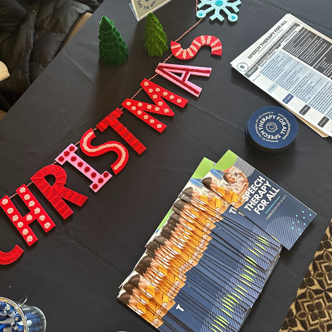 A black table with red and pink felt Christmas decorations spelling out 'CHRISTMAS,' green foam Christmas trees, blue snowflake, and a red-and-white candy cane decoration. Brochures titled 'Speech Therapy for All' are also on the table.