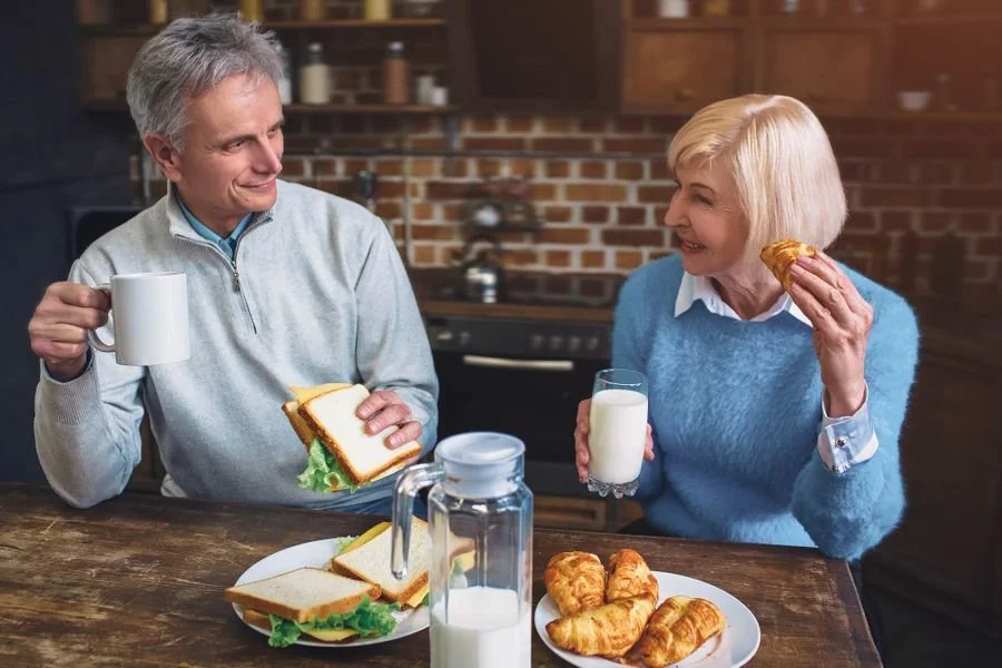 An elderly man and woman having breakfast together in a kitchen. The man is holding a coffee mug and a sandwich, while the woman is holding a glass of milk and a croissant. There are plates of sandwiches, croissants, a jug of milk, and some breakfast food on the table.