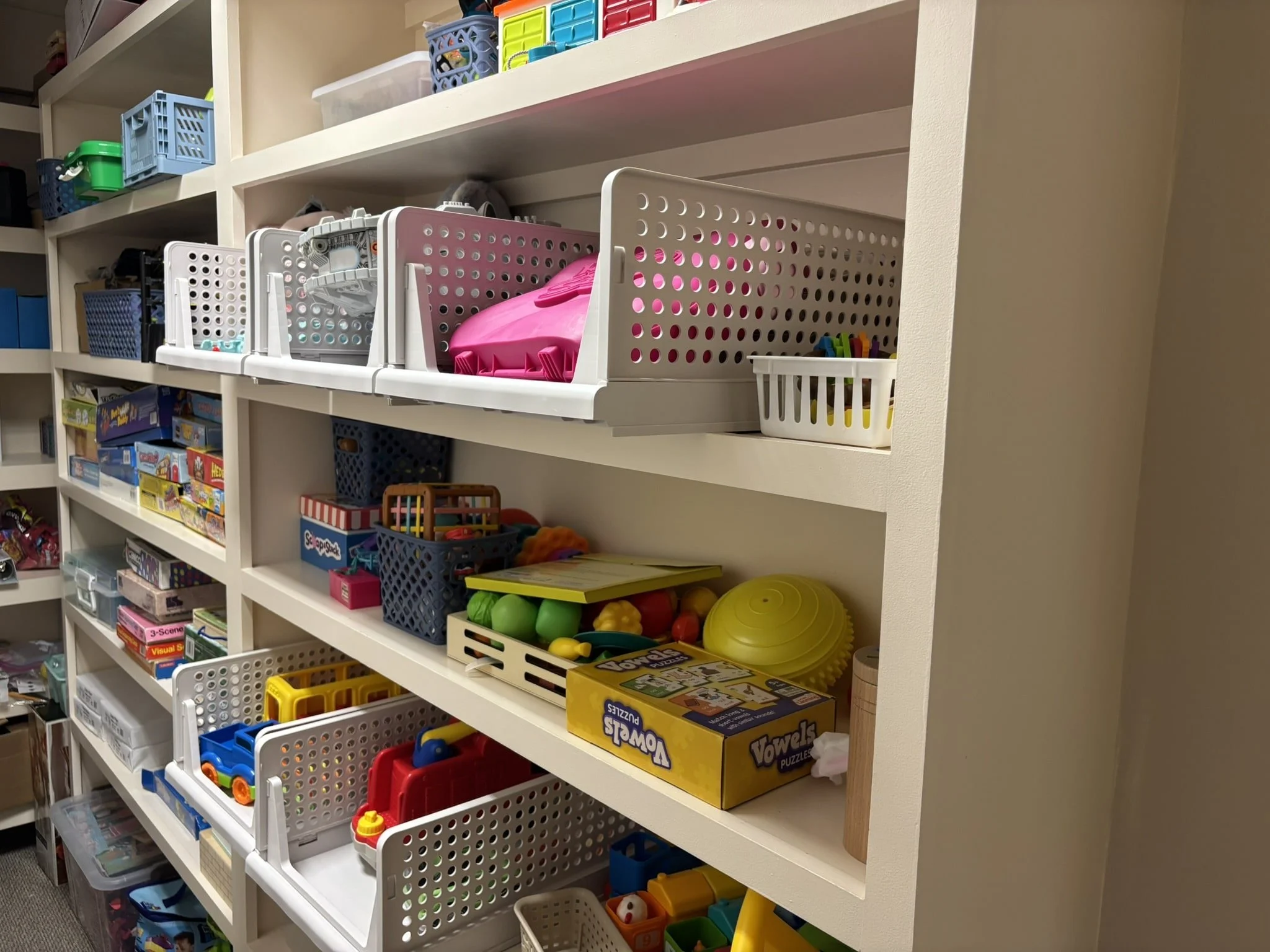 A white shelving unit filled with children's toys, games, and educational items organized in bins and on shelves.