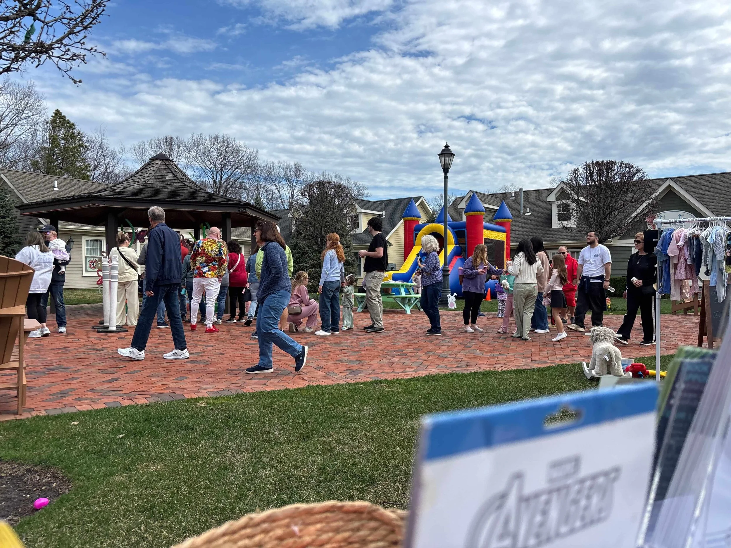 People gathered at an outdoor event in a park with a brick pathway, a colorful inflatable bounce house, and a pavilion with a dark shingled roof. Some are standing in line, walking, or talking, and there are children and adults present. A dog is sitting on the grass near a drink stand, and a clothes rack with hanging clothes is on the right.
