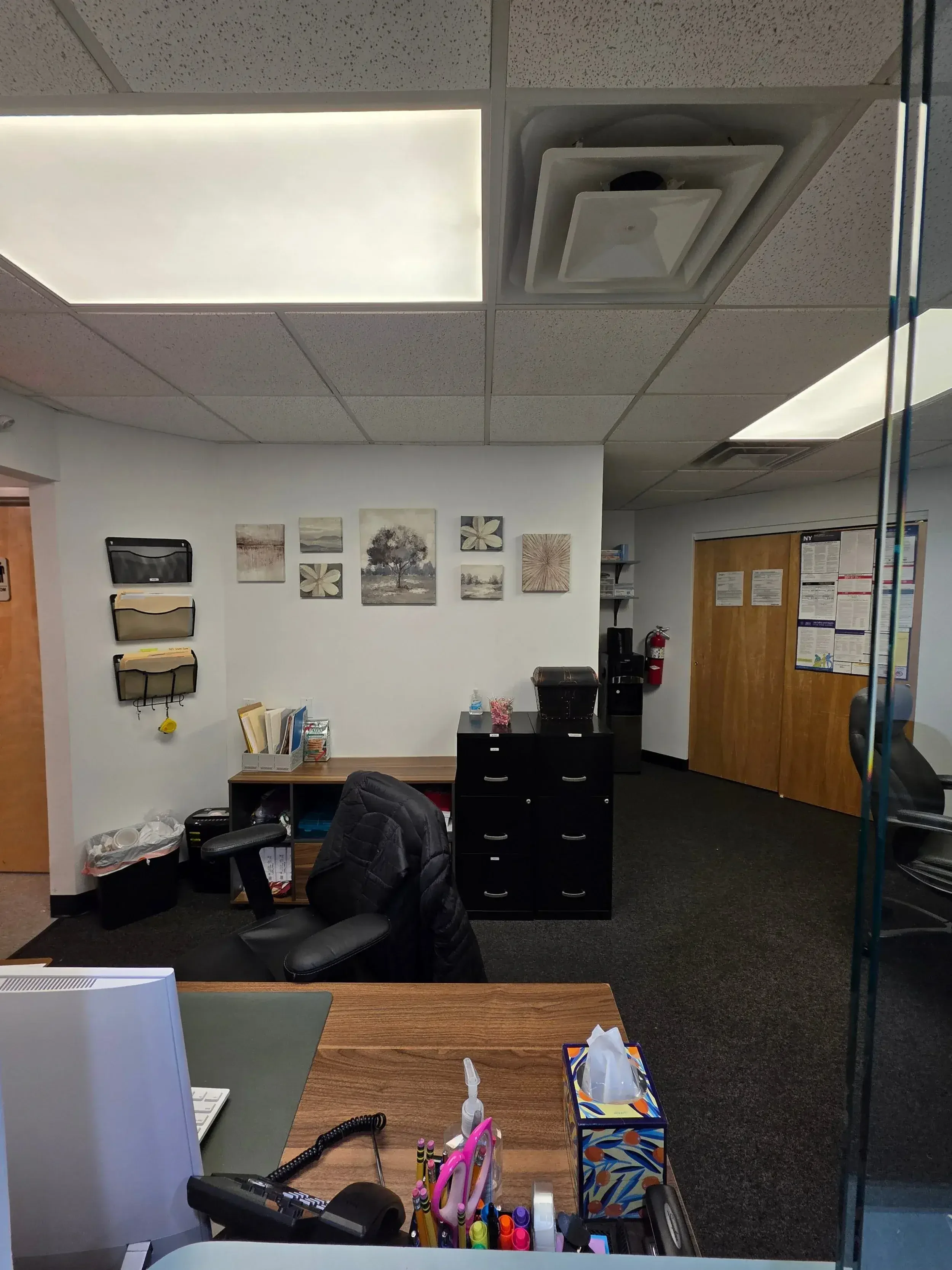 An office with a wooden desk, black office chair, and various office supplies, including a computer, pens, and tissues. The back wall has artwork and file organizers. There's a water cooler, fire extinguisher, and a closed wooden door.