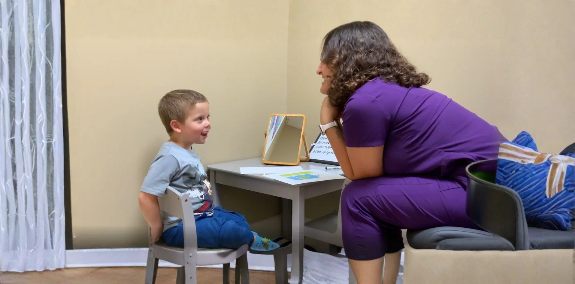 A young boy sitting on a small white chair facing a woman who is leaning over and smiling at him. They are in a room with beige walls, near a desk with a mirror and papers. The woman is wearing a purple outfit and the boy is wearing a gray t-shirt with a character on it and blue shorts.