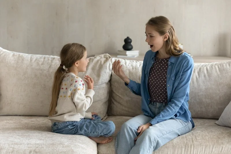 An adult woman and a young girl are sitting on a beige sofa, having an animated conversation, both appearing to be engaged and expressive.