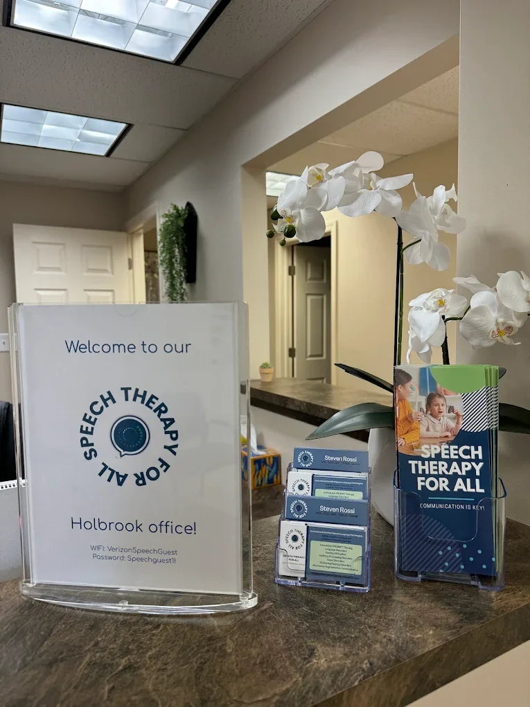 Reception desk at a speech therapy office with a sign that says "Welcome to our speech therapy all for all" and Wi-Fi information, as well as brochures and a decorative orchid plant.