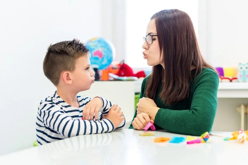 A woman and a young boy are sitting at a table facing each other, engaging in a serious conversation. The woman appears to be explaining something, while the boy listens attentively. In the background, there are toys, a globe, and other colorful items.