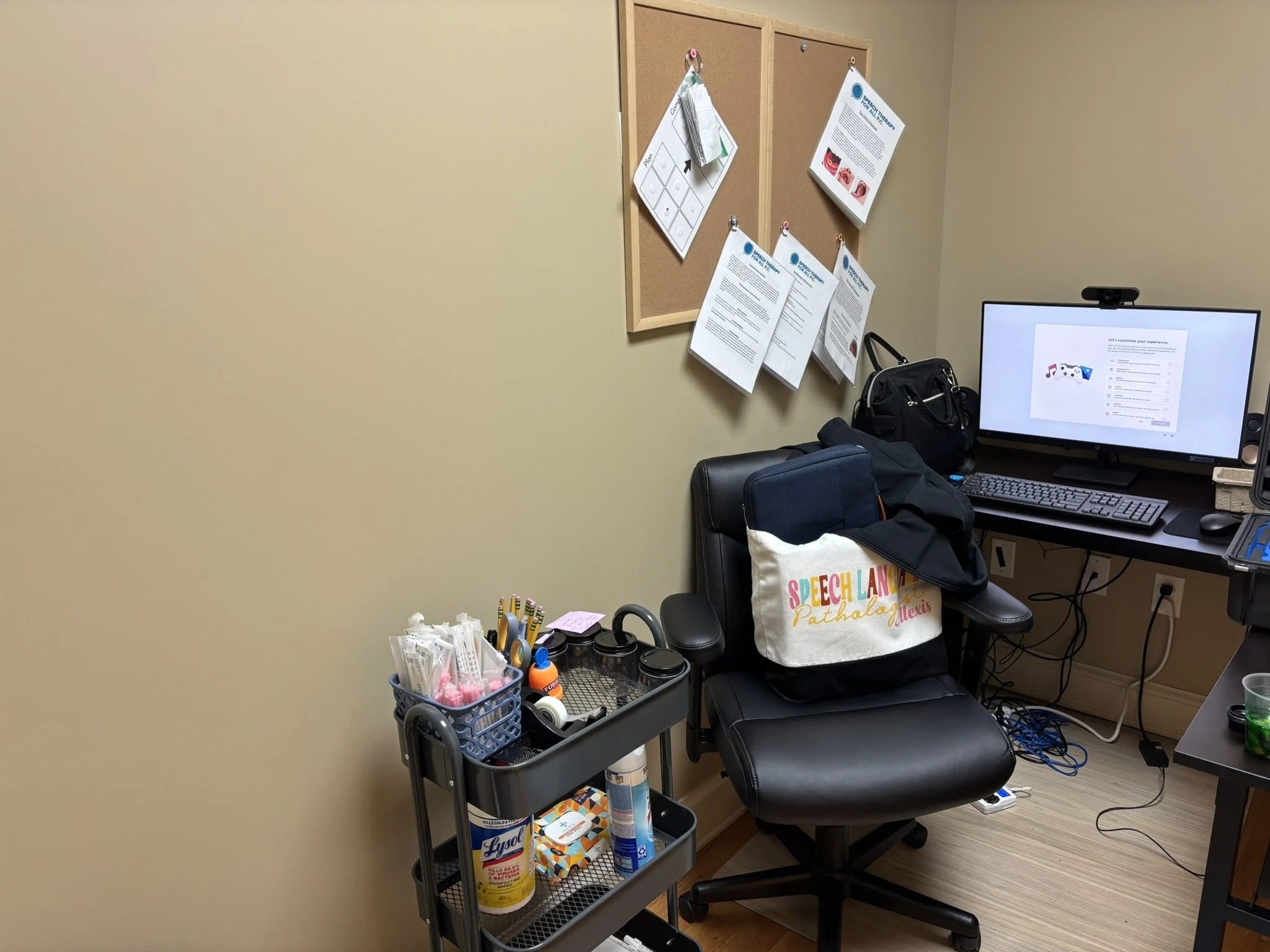An office corner with a black chair, a desk with a computer and a webcam, a bulletin board with papers pinned, a small cart with office supplies, and a tote bag that reads "Speech Land Pathology Alexis."