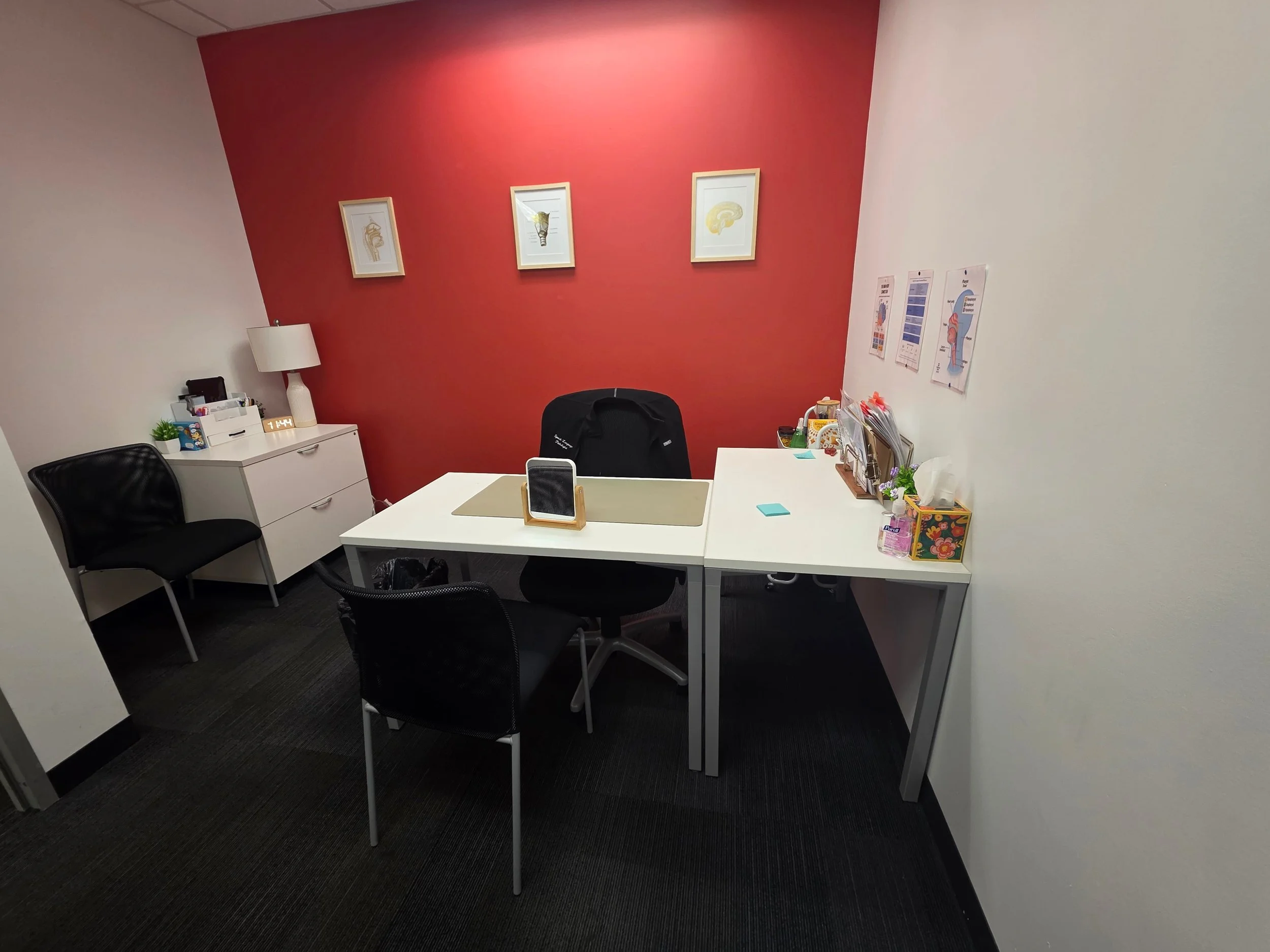 A small office with a red accent wall and three framed anatomical prints. Contains a white desk with a black office chair, and a bedside table with a white lamp, green plant, and office supplies. A black guest chair is in front of the desk.