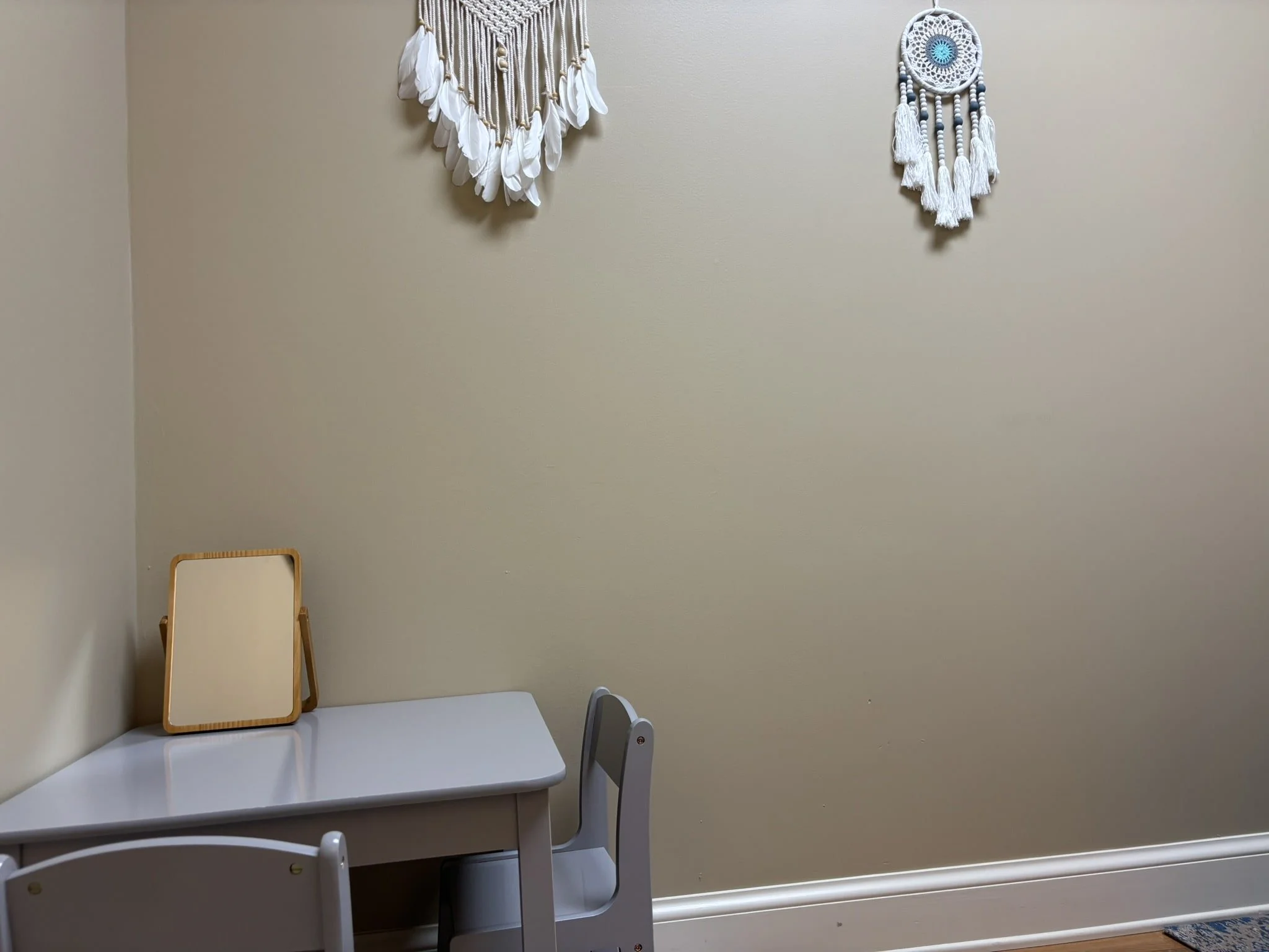 A small white table with a mirror on top, two white chairs, and two bohemian wall hangings on a beige wall.