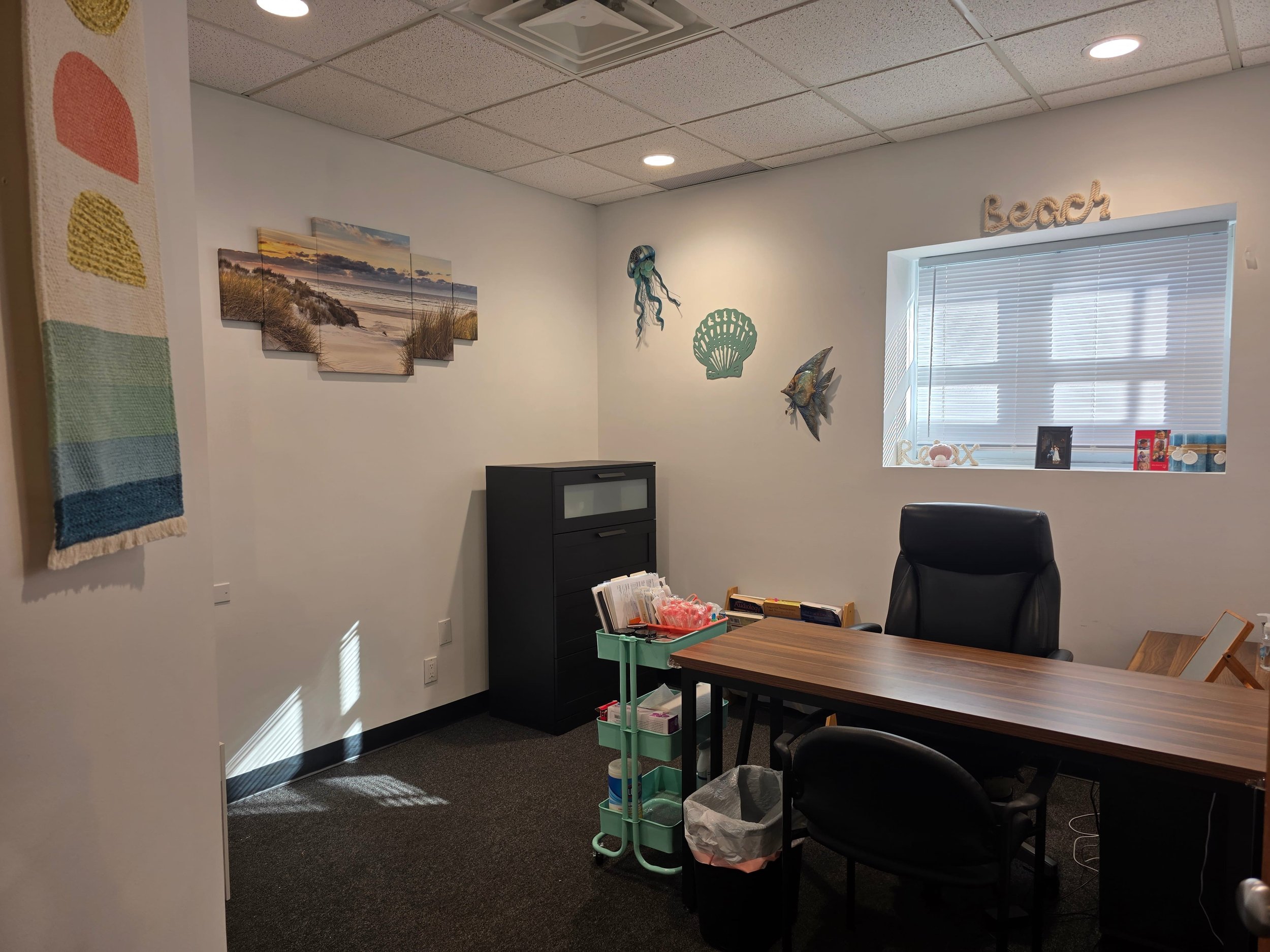 Empty office with a wooden desk, black office chairs, wall decorations including a mural of a beach, coral, a fish, and a jellyfish, and a window with blinds.