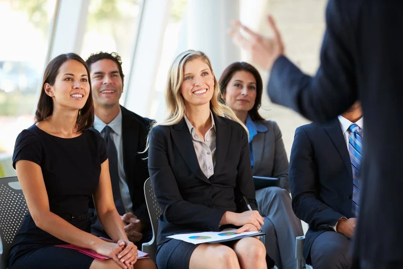 A diverse group of business professionals sitting in a meeting room listening to a presentation, with a woman in front speaking.