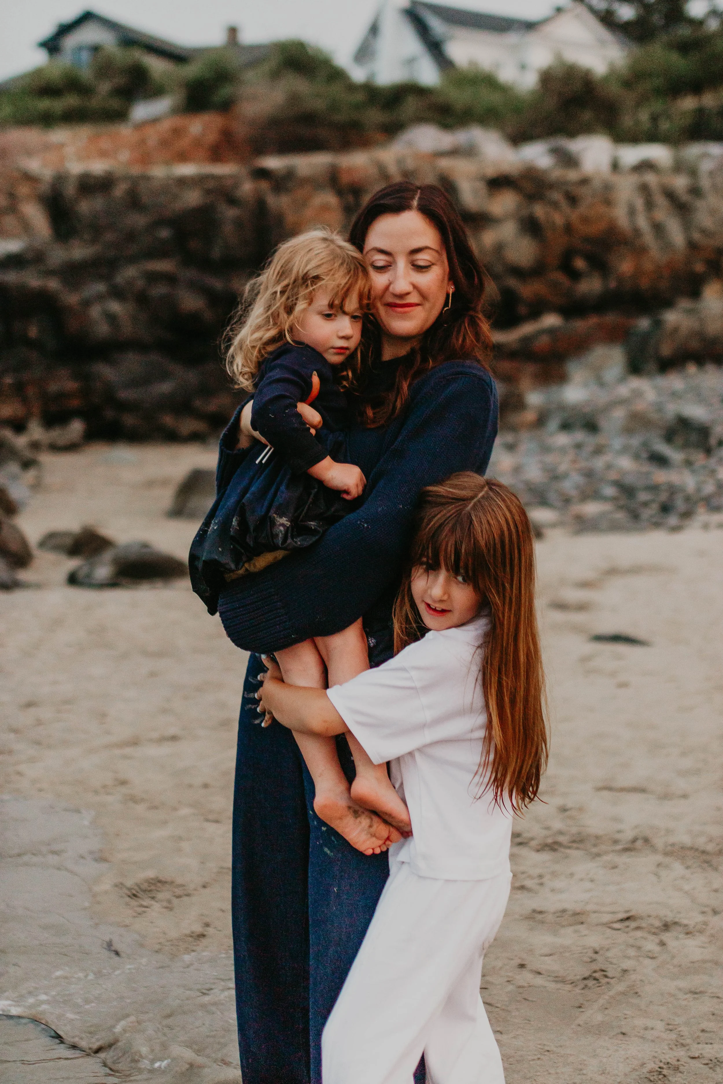 A woman with long dark hair hugging two young girls at the beach. One girl has blonde curly hair and is wearing a dark jacket, while the other girl has long straight red hair and is wearing a white shirt. There are rocks and houses in the background.