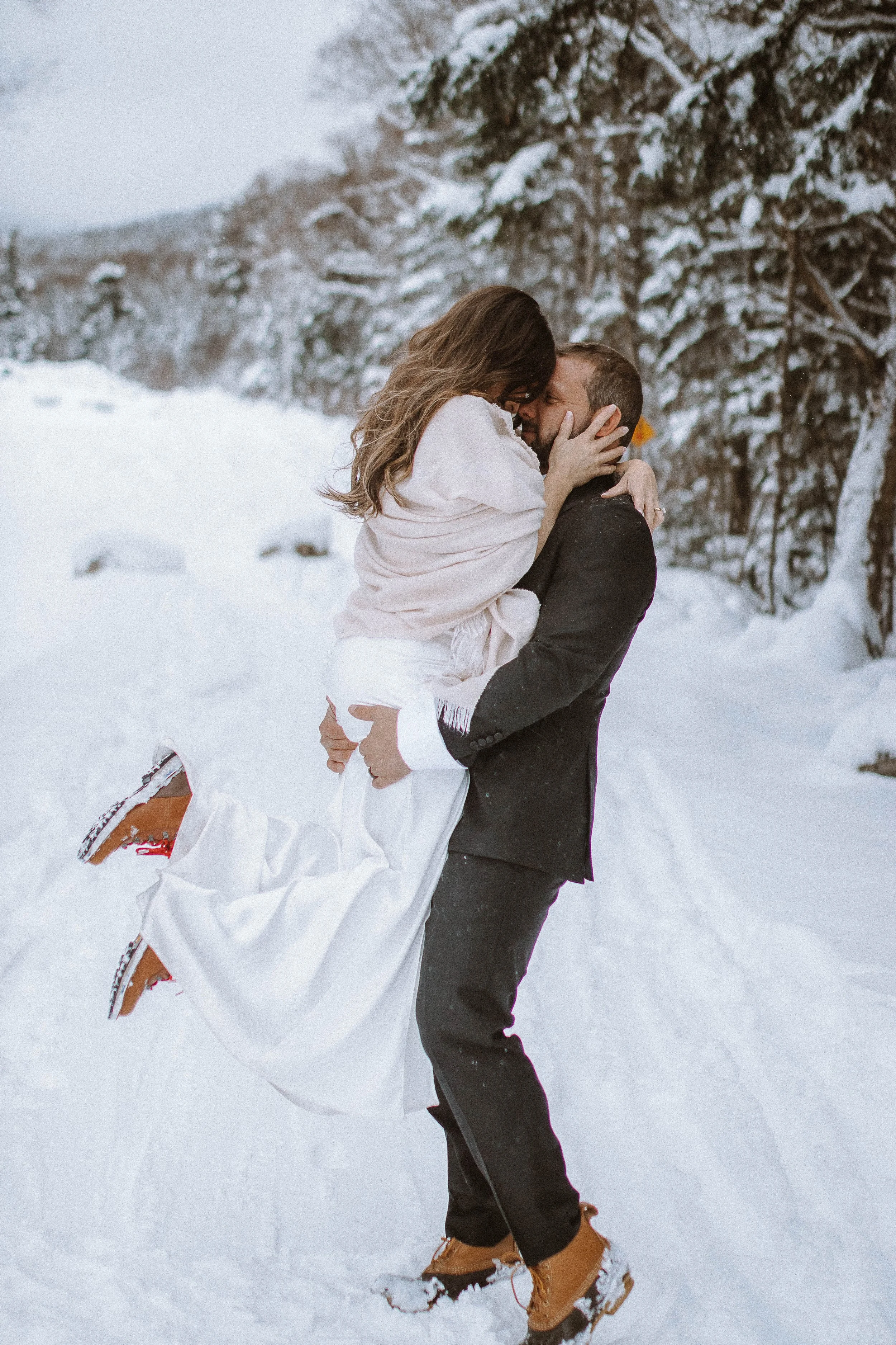 A man in a black suit lifts a woman in a white dress in the snow, surrounded by snow-covered trees, in a wintery outdoor setting.