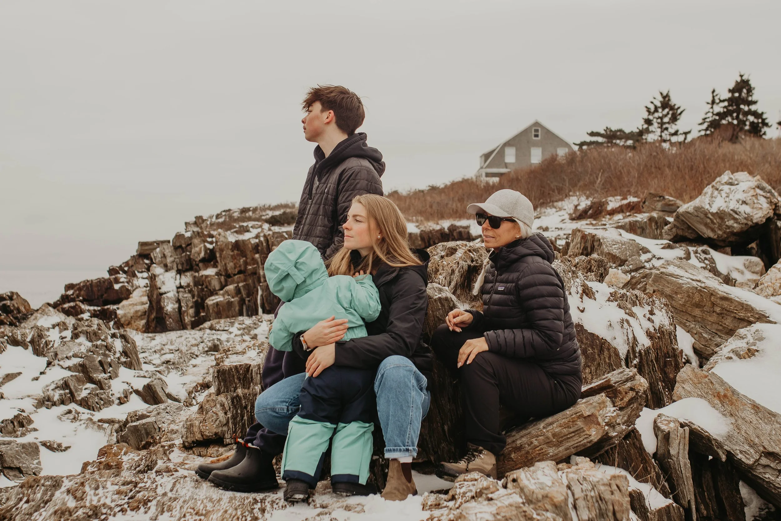 Four people on a snowy rocky shore with a house in the background. One young woman with a child hugging her, another older woman wearing sunglasses and a cap, and a young man standing behind them.