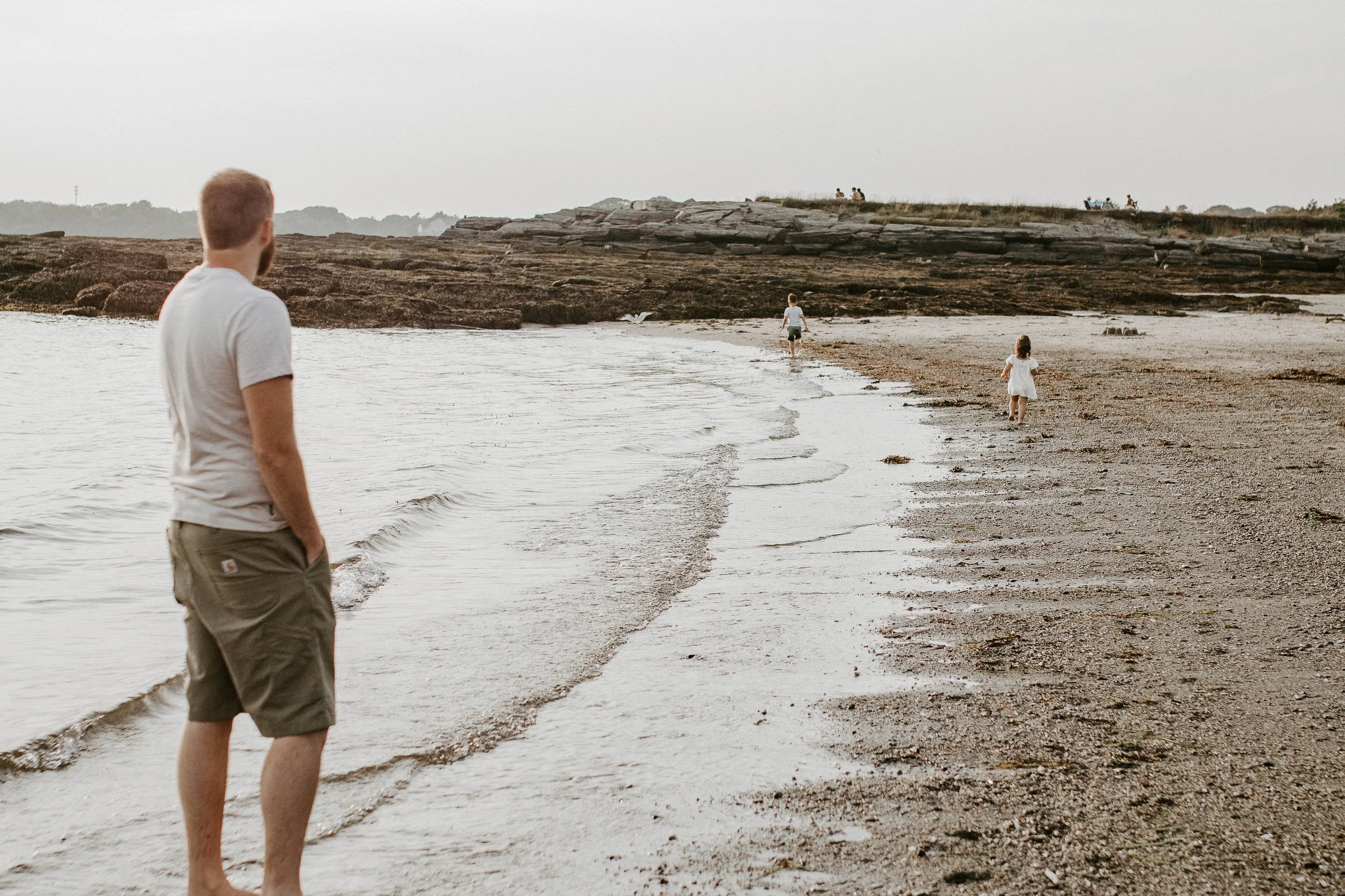 A man standing on the sandy shore watching children playing near the water. The shoreline extends into the distance with rocks and a few people sitting on top of a rocky outcrop.