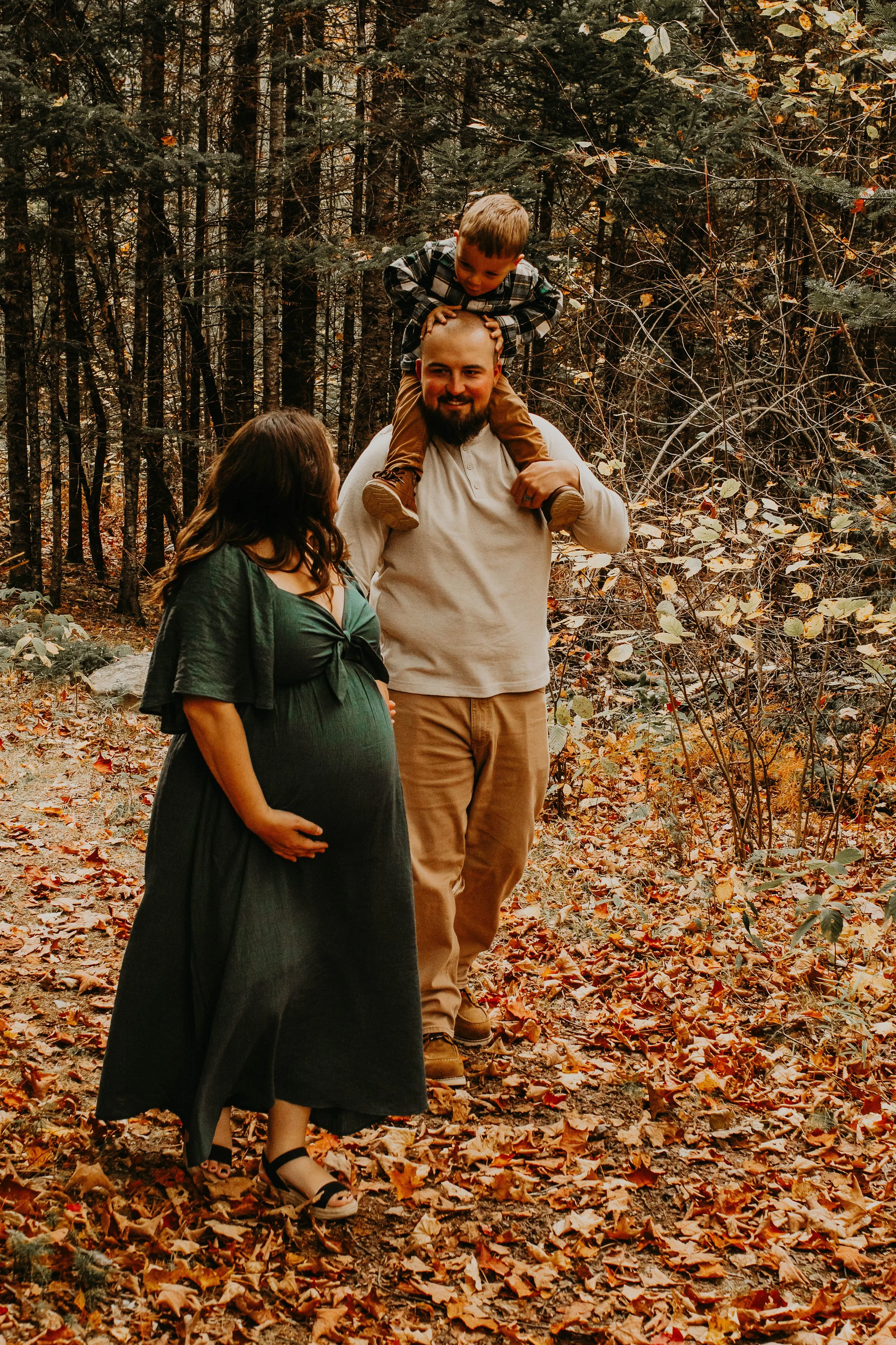 Family of three celebrating outdoors in a forest during autumn, with fallen leaves on the ground and trees in the background. The pregnant woman is wearing a dark dress and sandals, the man is carrying a young child on his shoulders, and the woman is gently holding her pregnant belly.