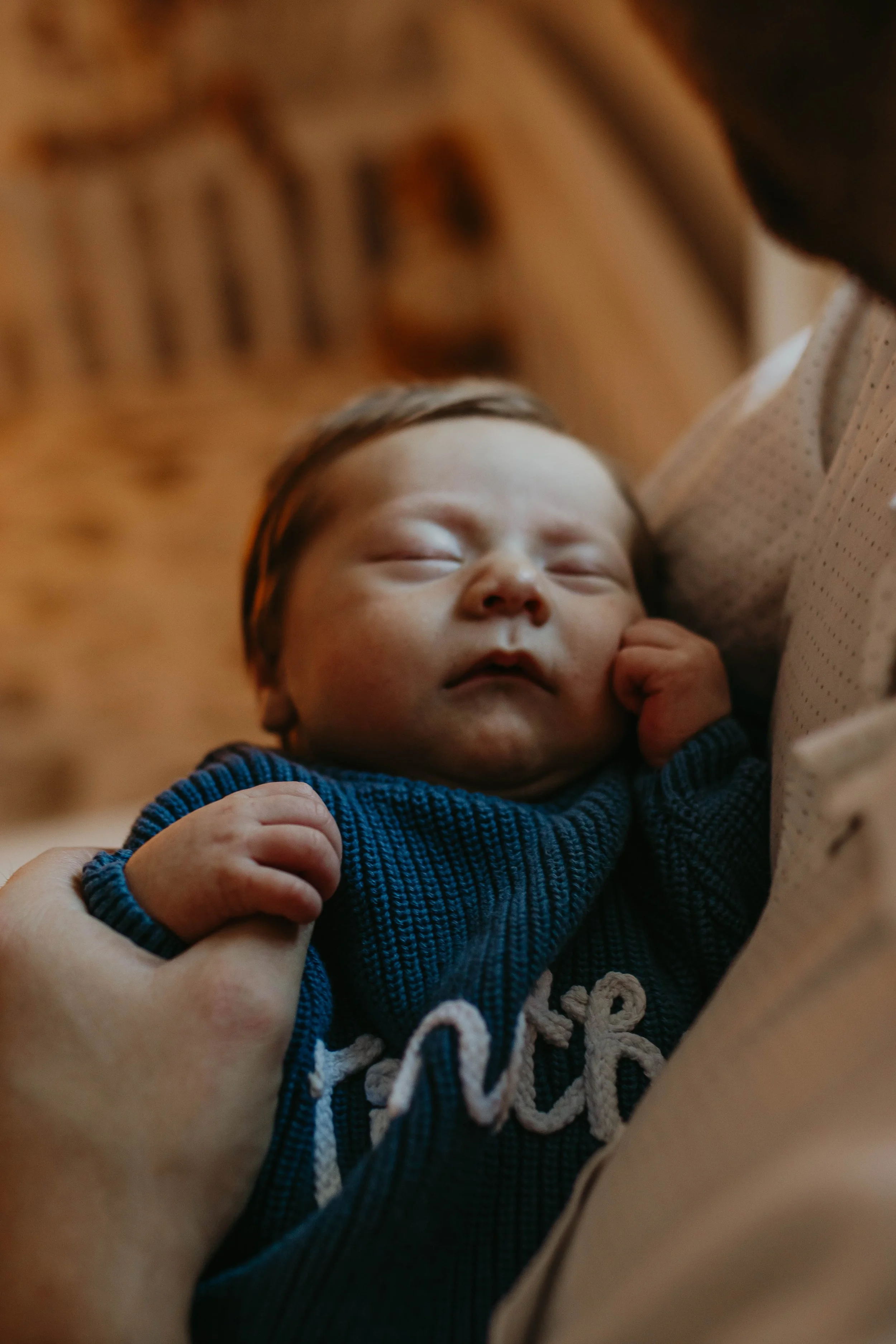 Close-up of a sleeping newborn baby being gently held, wearing a dark blue sweater with white embroidery.