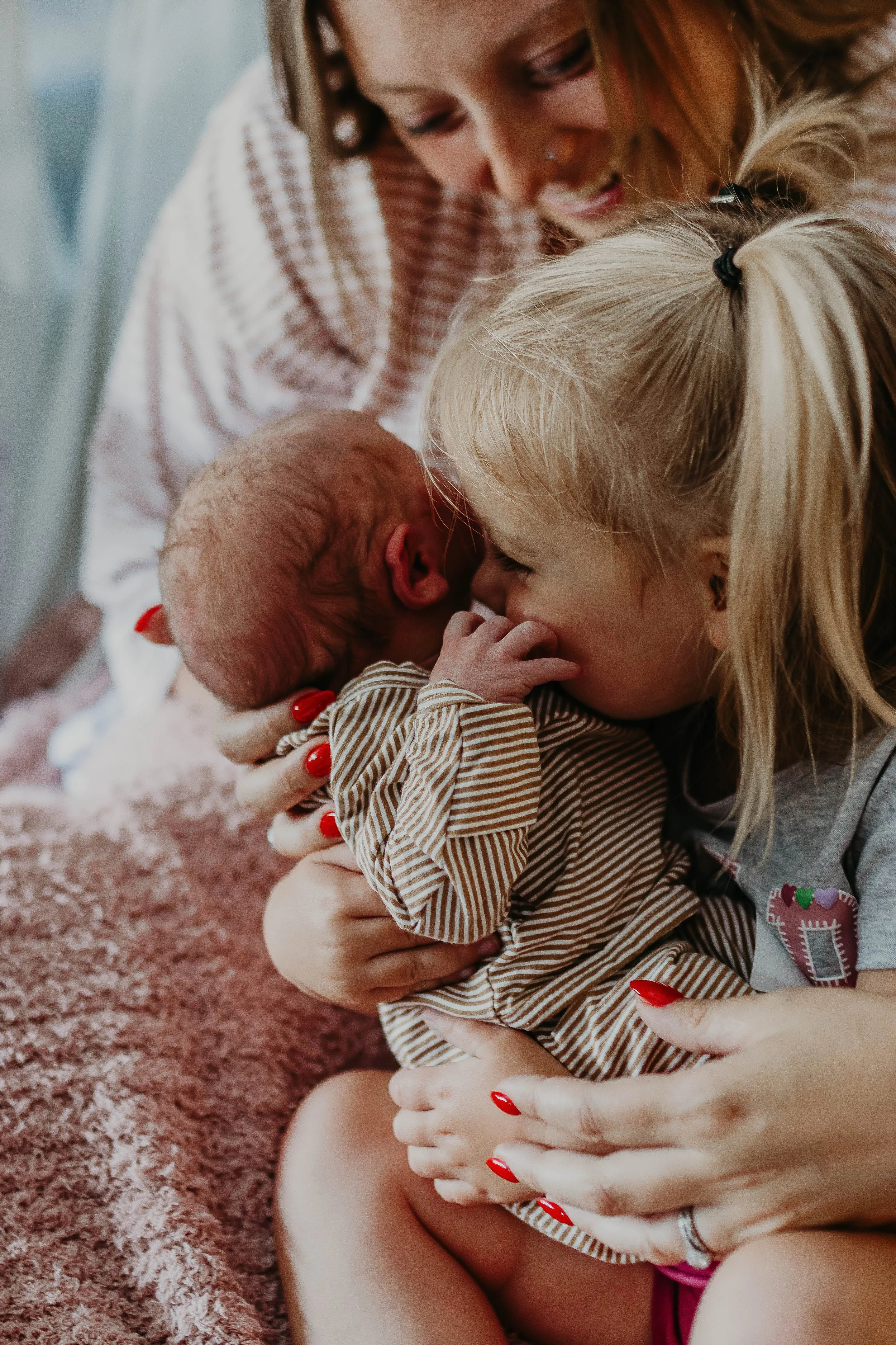 A woman with red nails holding a young blonde girl in a striped shirt, both smiling. The girl is holding a small, bald baby with closed eyes, cradled in the woman's arms. They are sitting on a pink, fuzzy carpet.