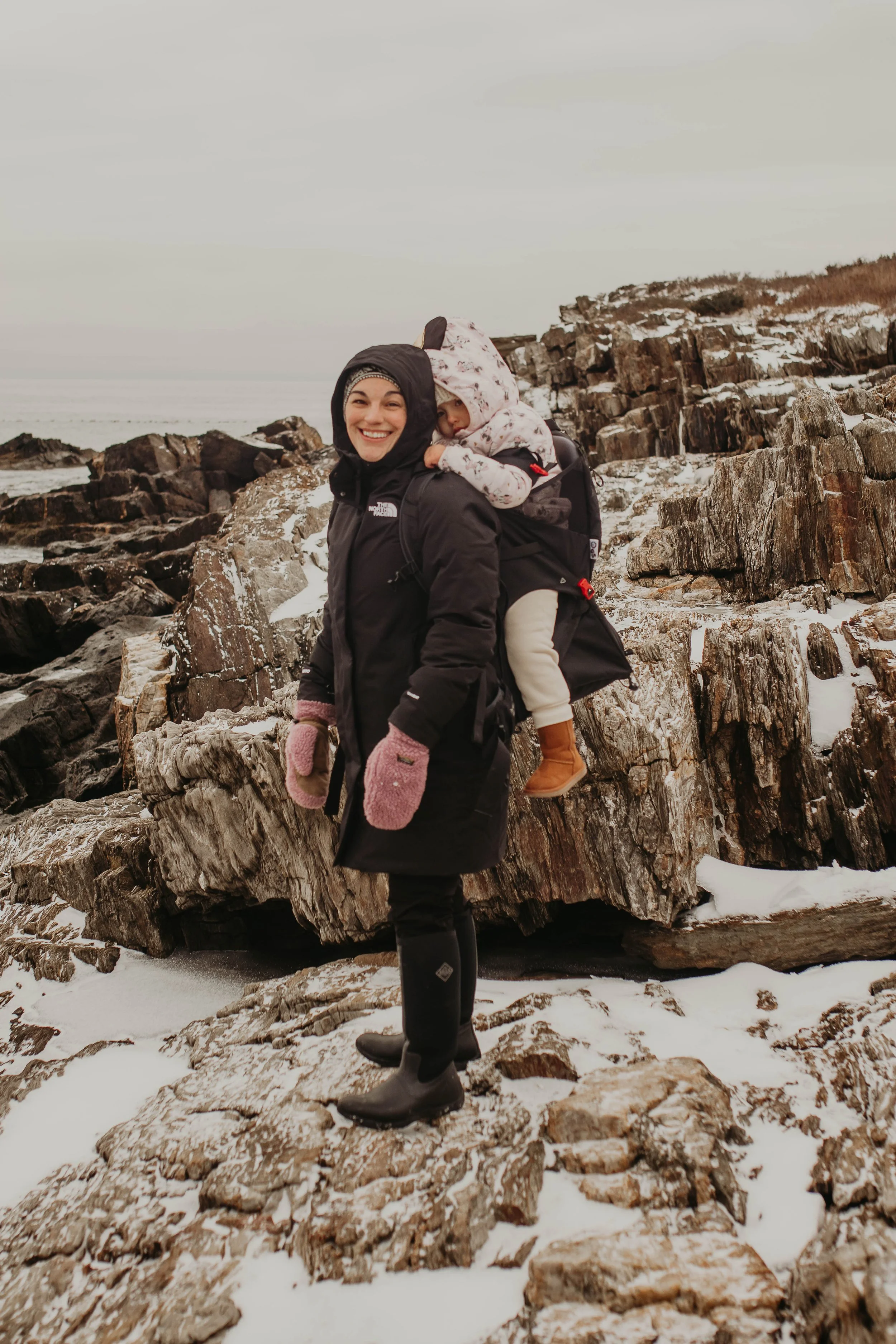 Woman smiling while carrying a young child on her back on a rocky, snowy coastline.