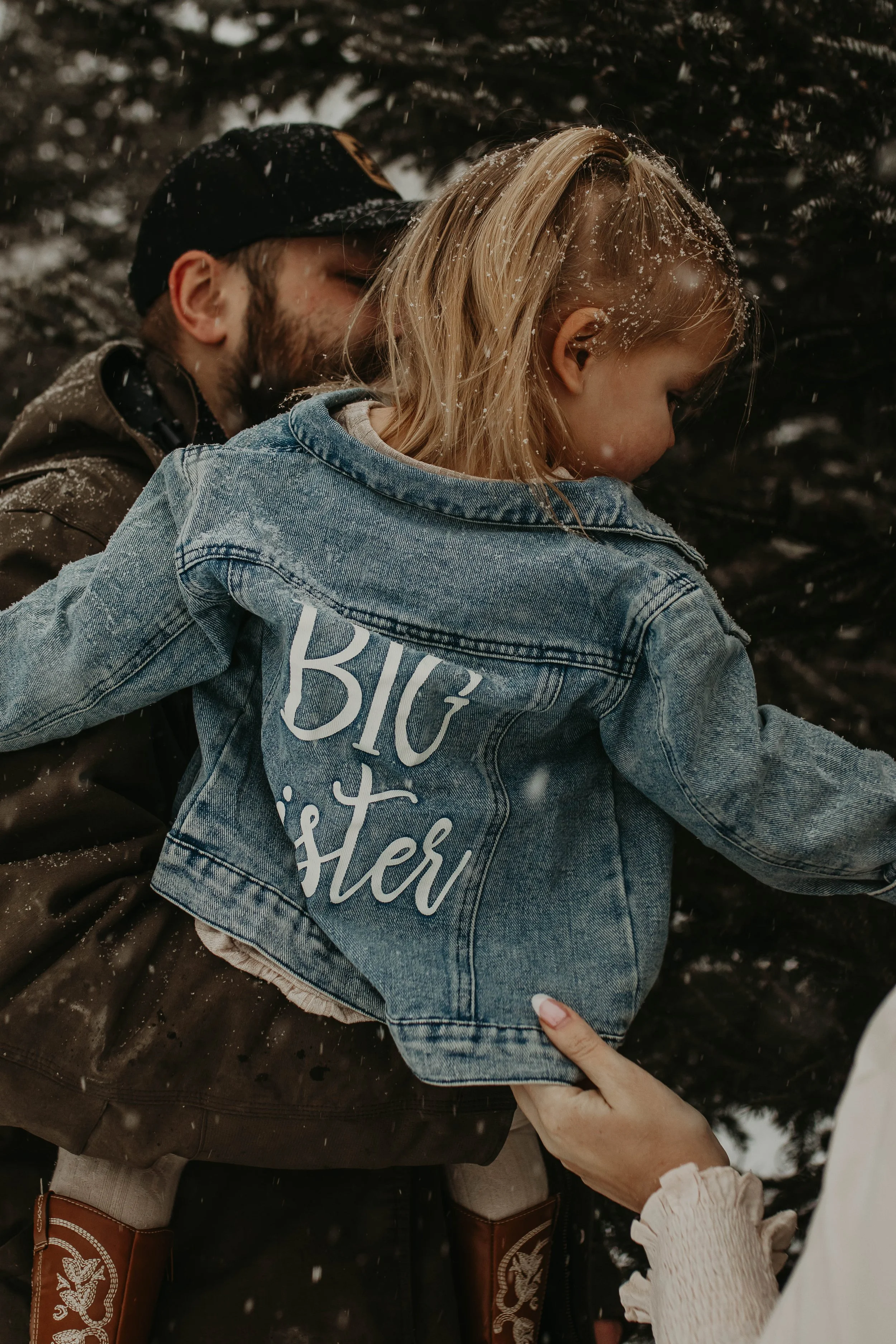 A blonde girl wearing a denim jacket with 'Big Sister' written on the back, being carried by a man in a brown jacket and black cap, outdoors with snow and trees in the background.