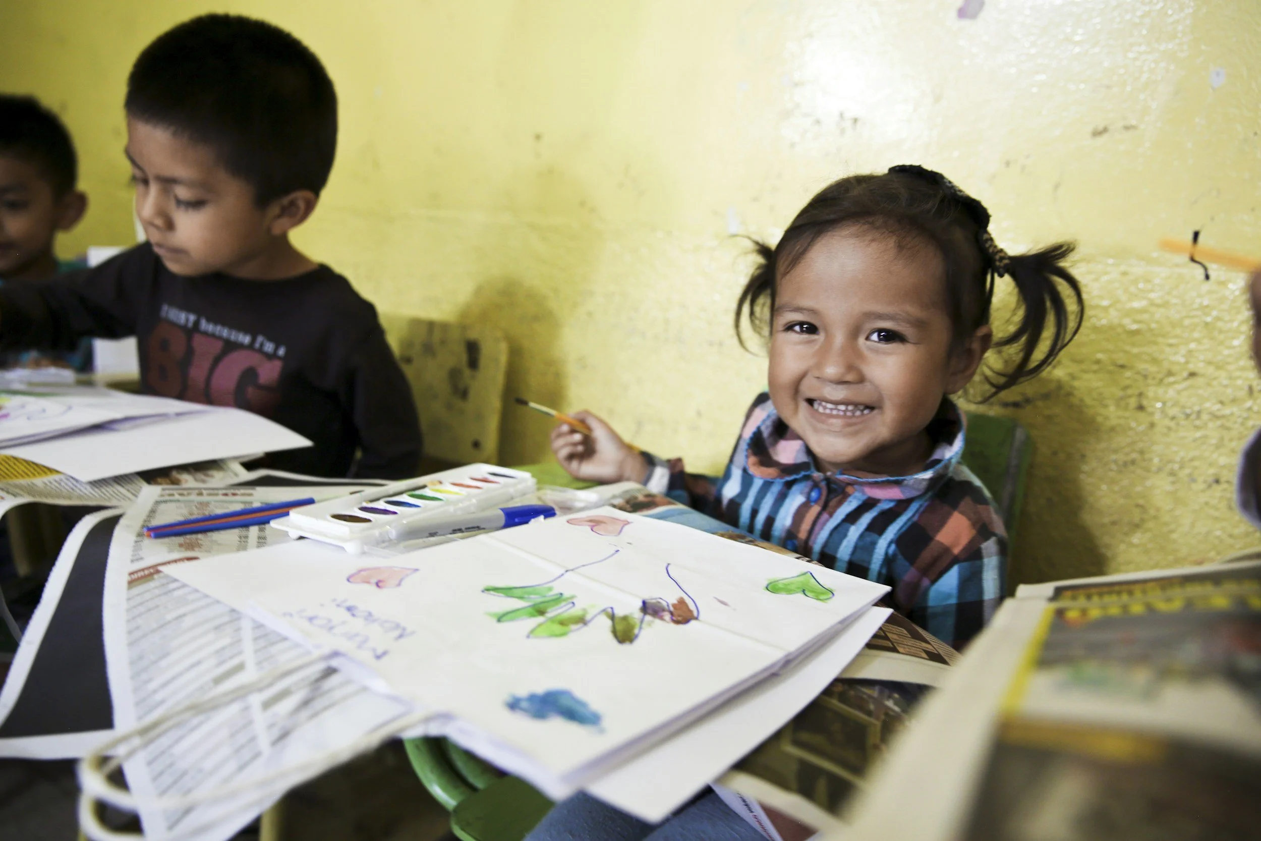 Smiling young girl holding a paintbrush while drawing with classmates at a classroom table