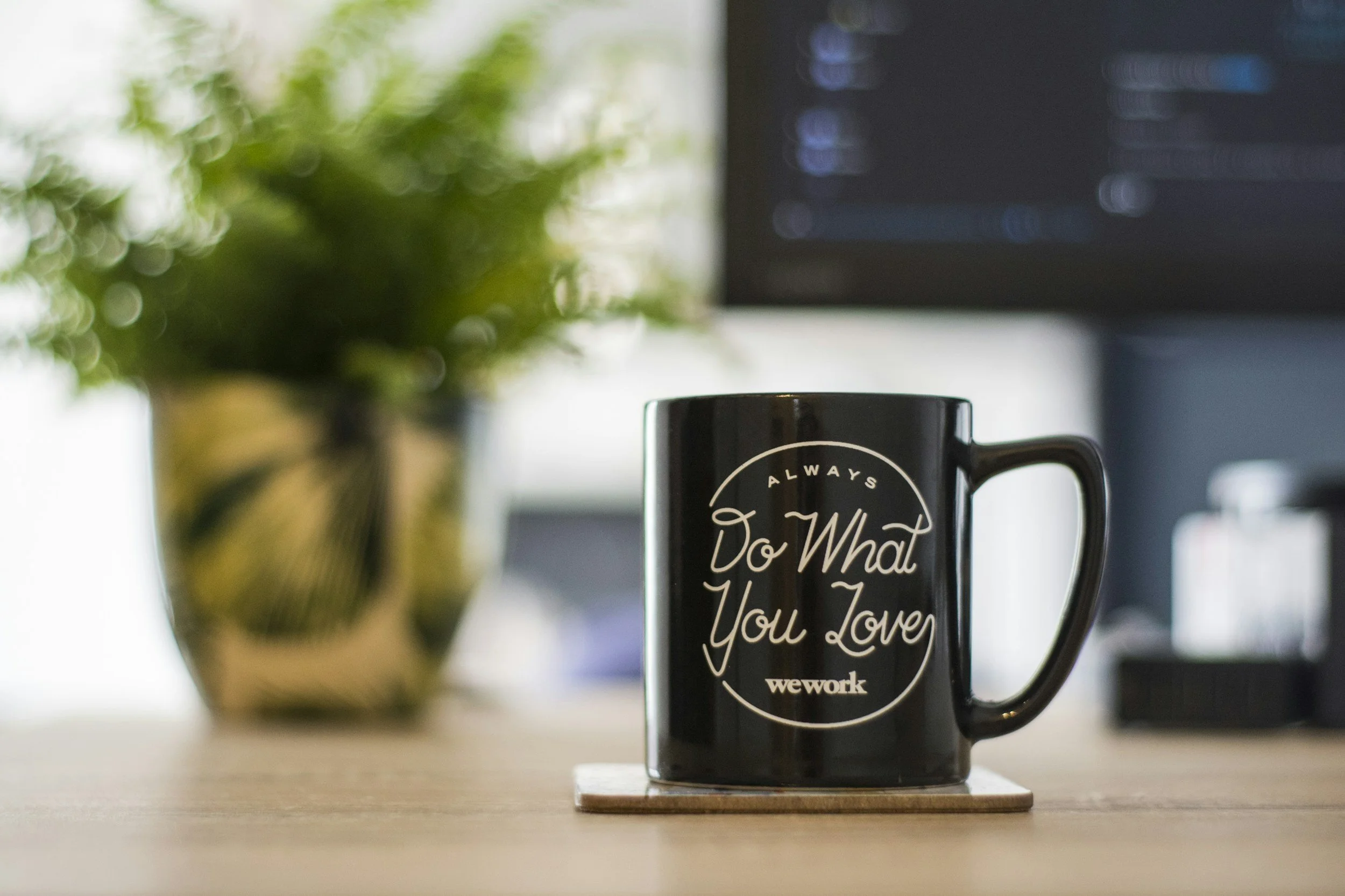 Black WeWork coffee mug with motivational quote on a wooden desk beside a blurred potted plant and computer monitor