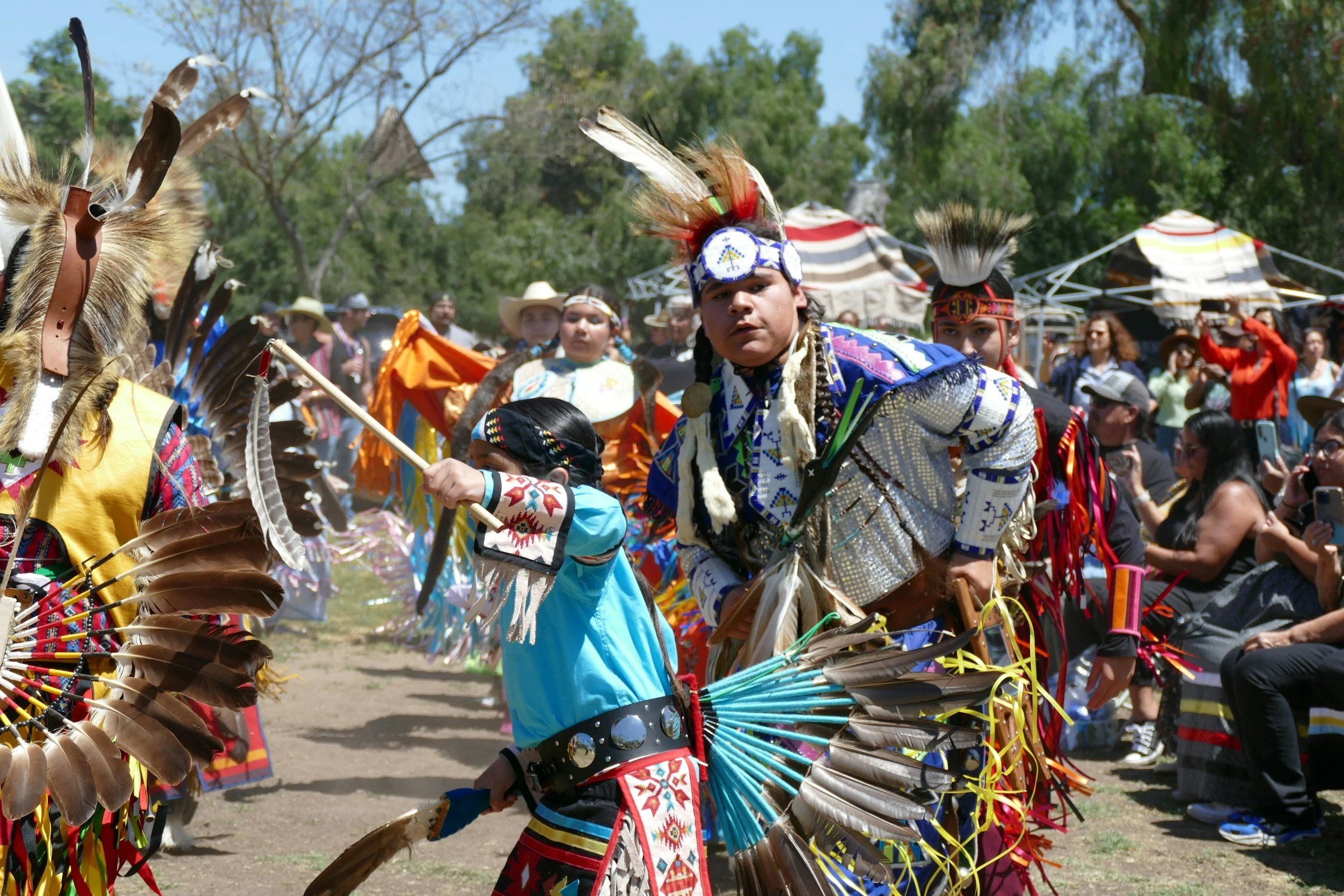 Native American dancers in colorful feather regalia perform at an outdoor powwow as spectators watch.