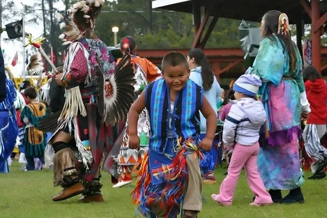 Children and adults in colorful traditional Indigenous regalia dancing at an outdoor cultural celebration.