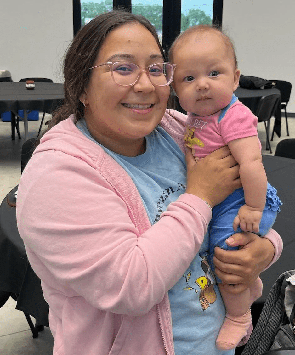Smiling woman wearing glasses holds a baby in a pink shirt and blue pants indoors.