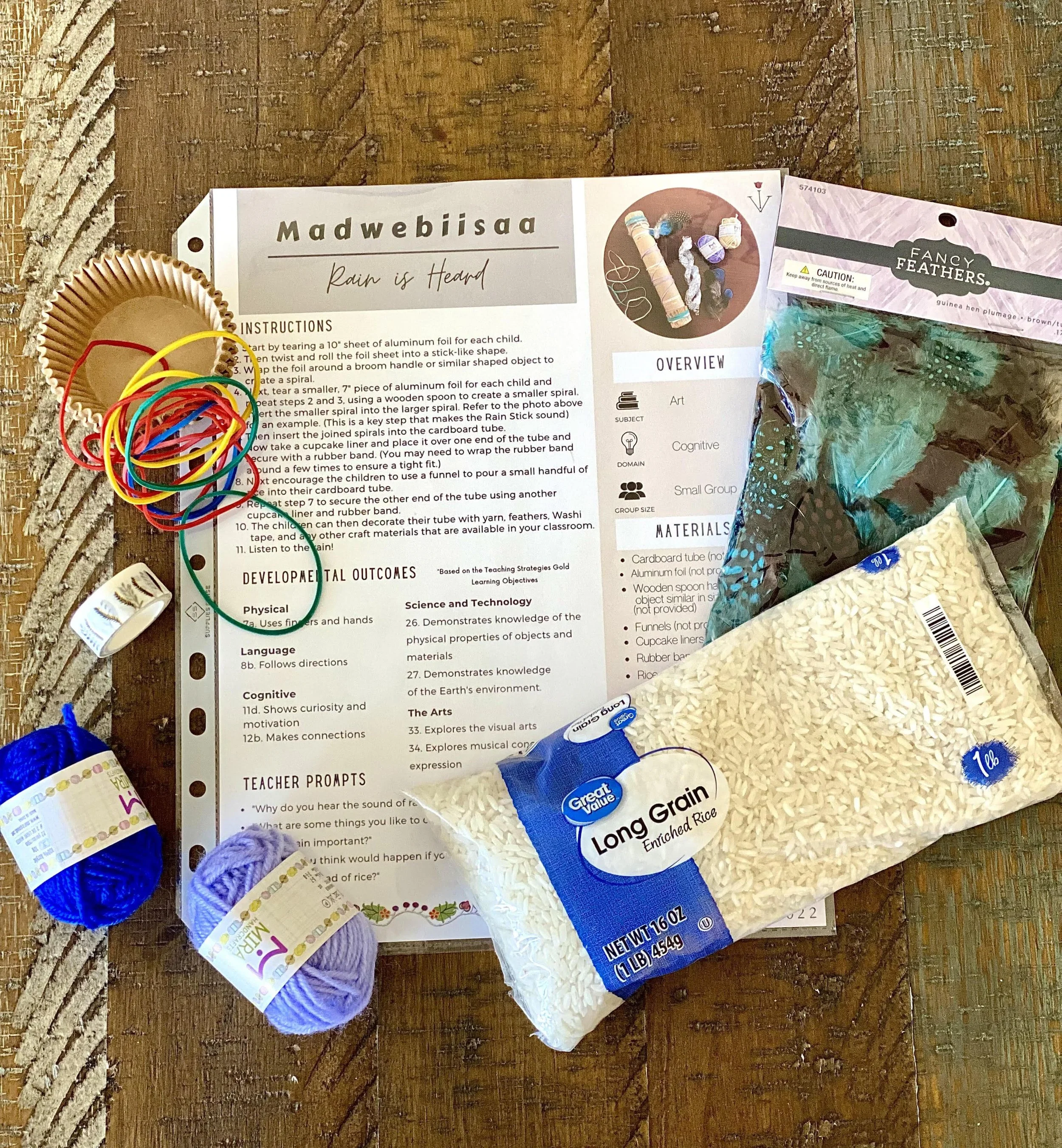 Rain stick craft activity supplies, including instructions, rice, feathers, yarn, rubber bands, cupcake liners, and craft materials on a wooden table