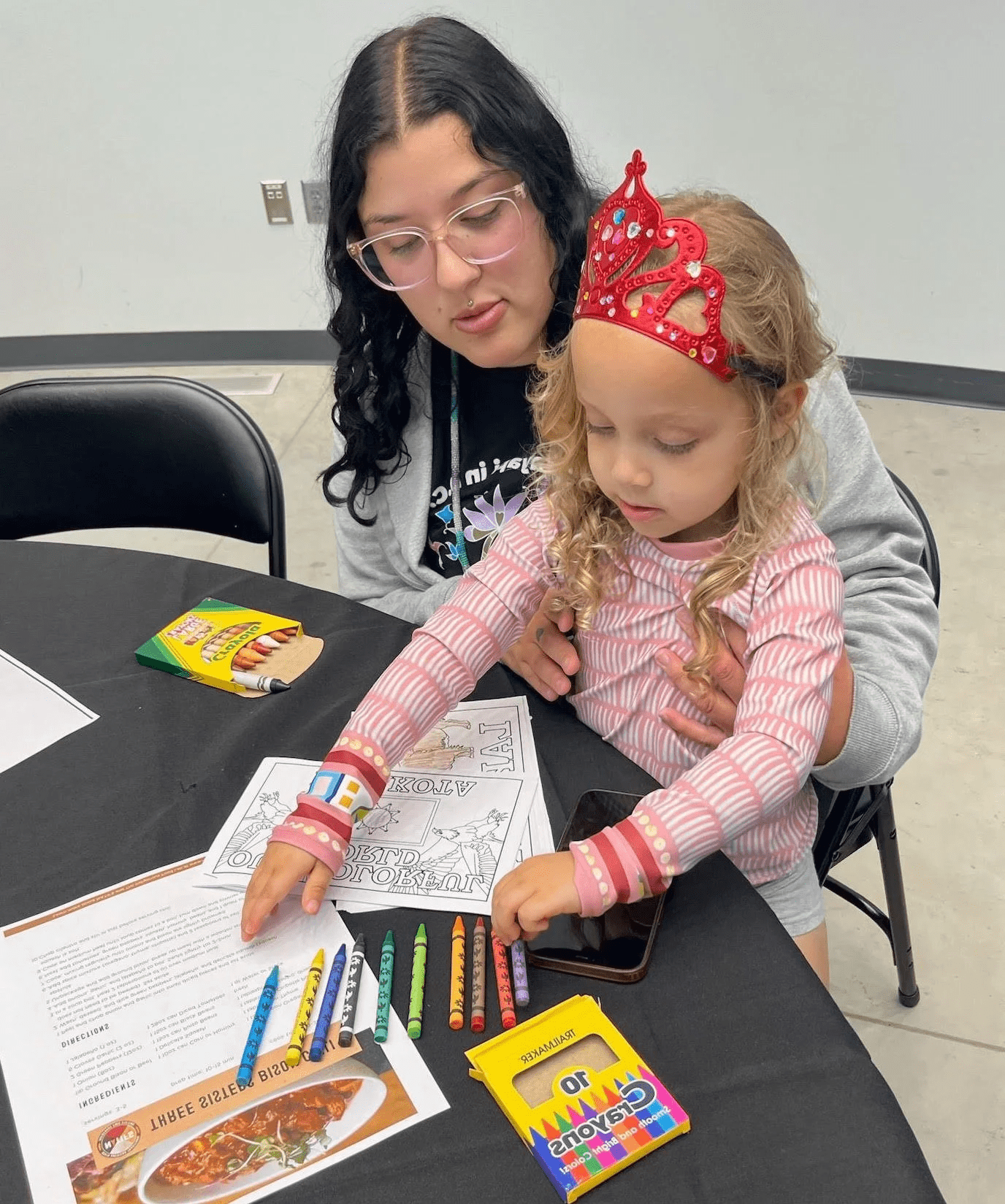 Adult helping a young girl wearing a red jeweled crown color worksheets at a table with crayons