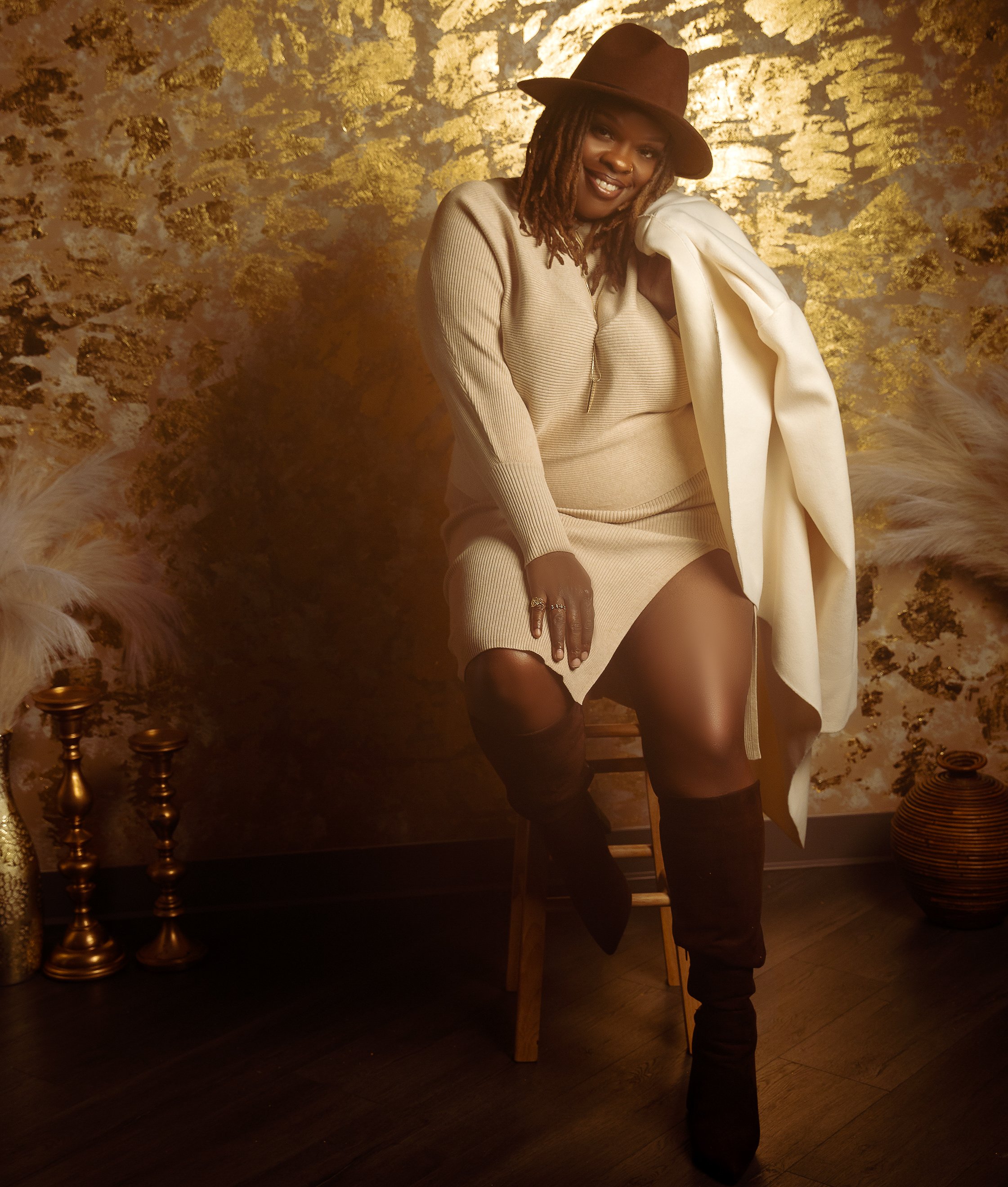 A woman with dreadlocks wearing a brown hat and beige outfit, sitting on a wooden stool in a room with gold and cream decor, including gold candlesticks, a gold vase with feathers, and a gold-brown woven vase.