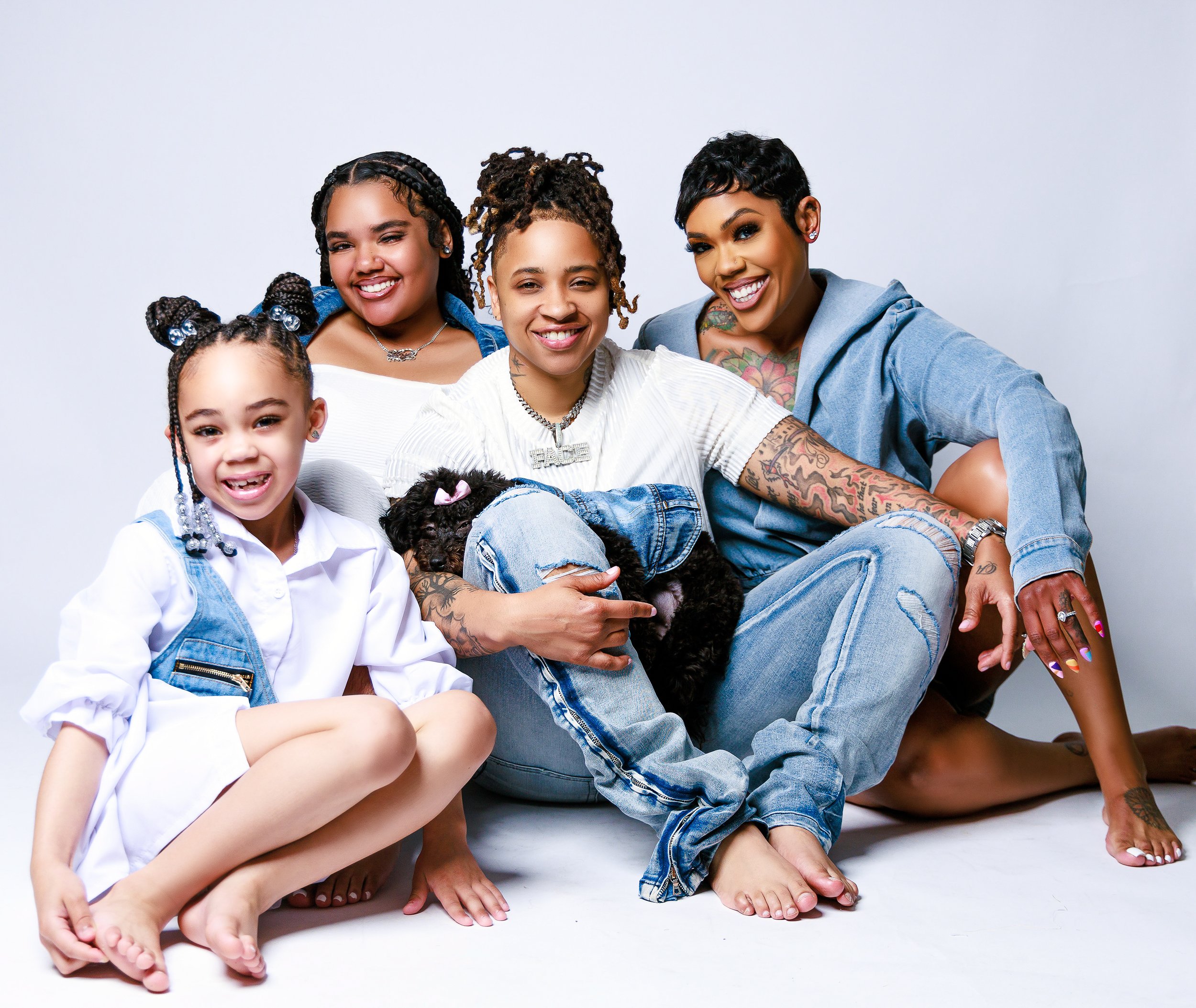 A diverse group of four women and one young girl sitting together on the floor, smiling, with a small black puppy.