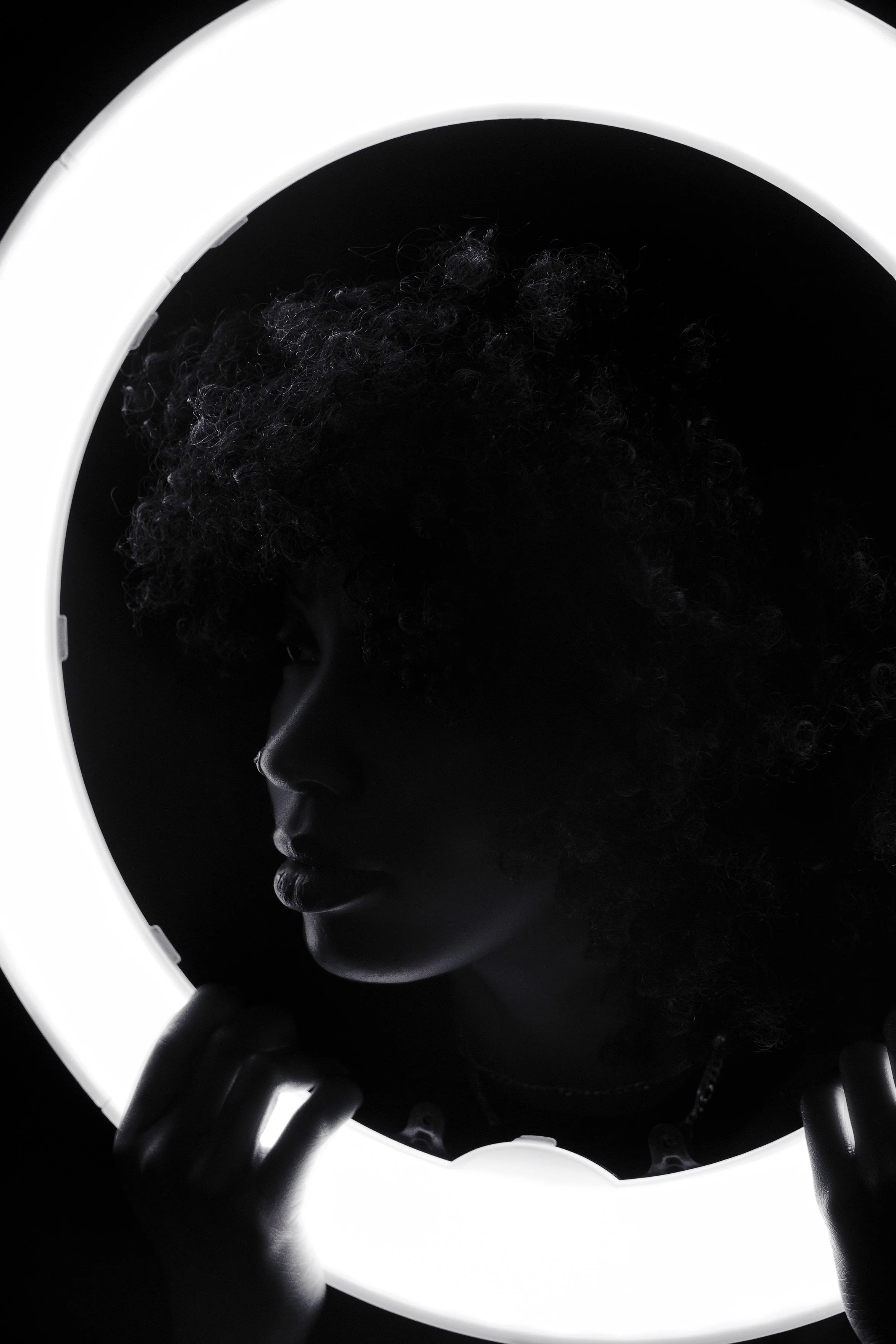 Silhouette of a person with curly hair holding a round, illuminated mirror in a dark setting.