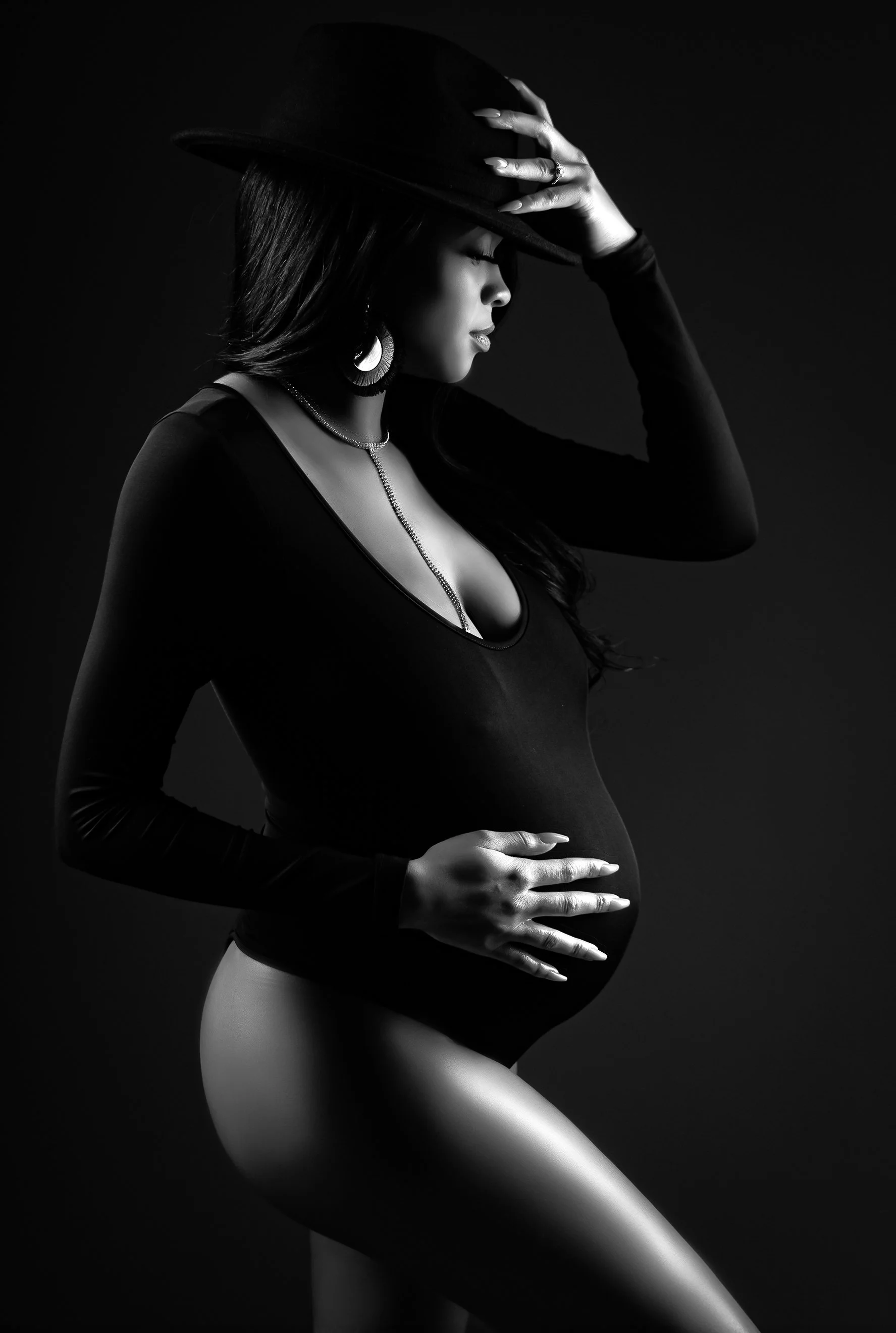 A black-and-white portrait of a pregnant woman wearing a hat, long sleeves, and jewelry, with her eyes closed and hand on her belly.