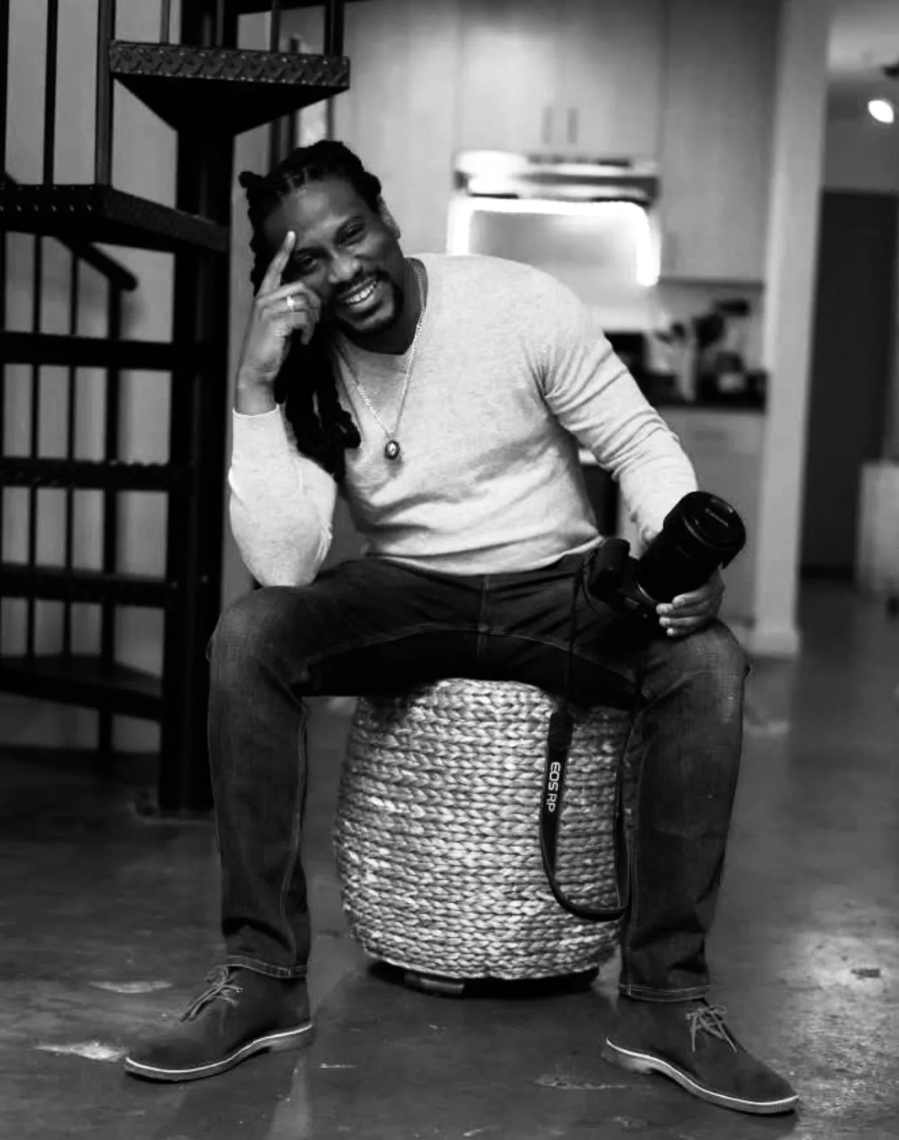 A man with dreadlocks sitting on a woven stool in a room, holding a camera in his right hand, smiling, wearing a light-colored long-sleeve shirt, dark jeans, and boots, with a background of shelves and kitchen area.