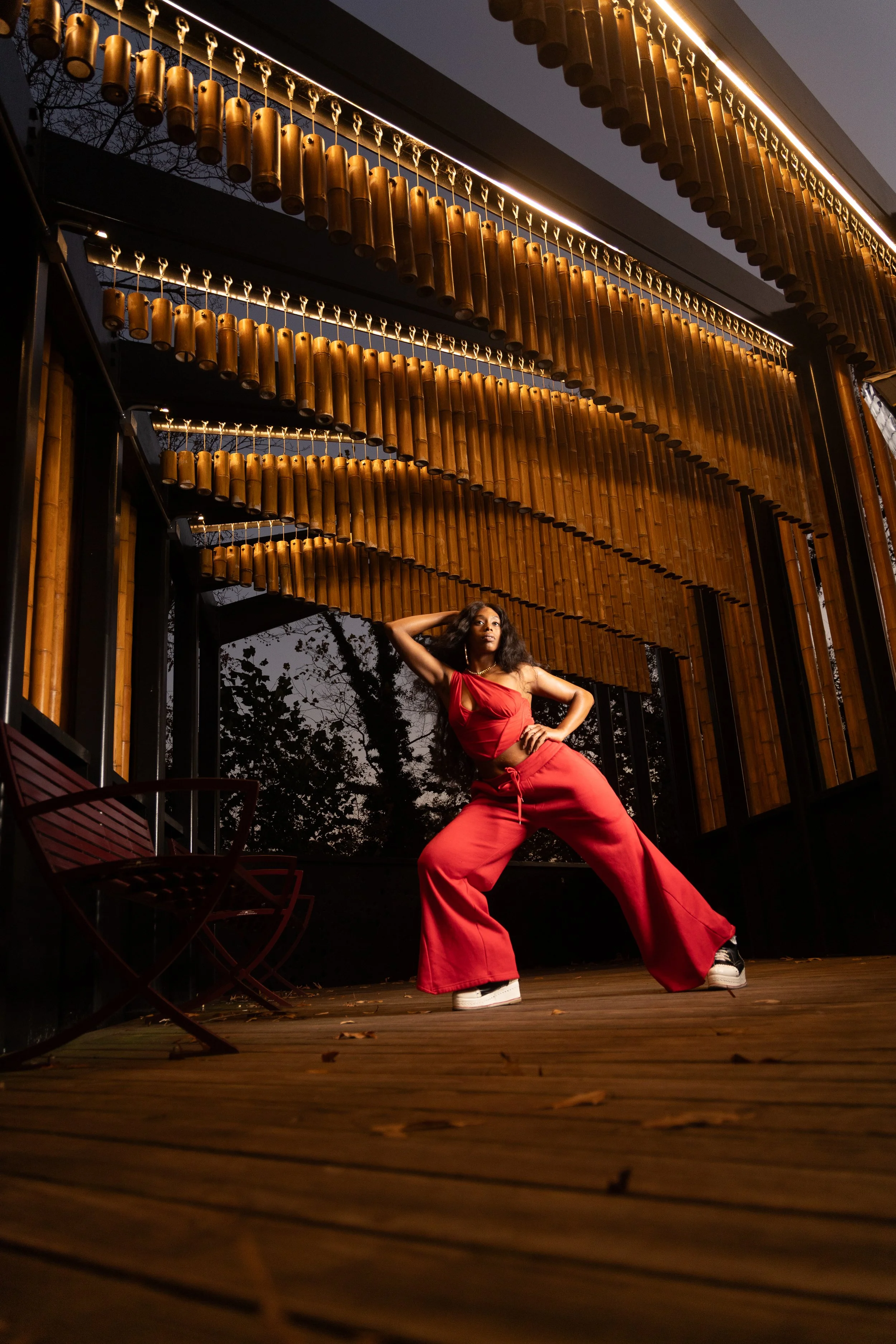A woman in a red outfit poses confidently on a wooden deck, with a modern building featuring hanging cylindrical decor behind her, during dusk.