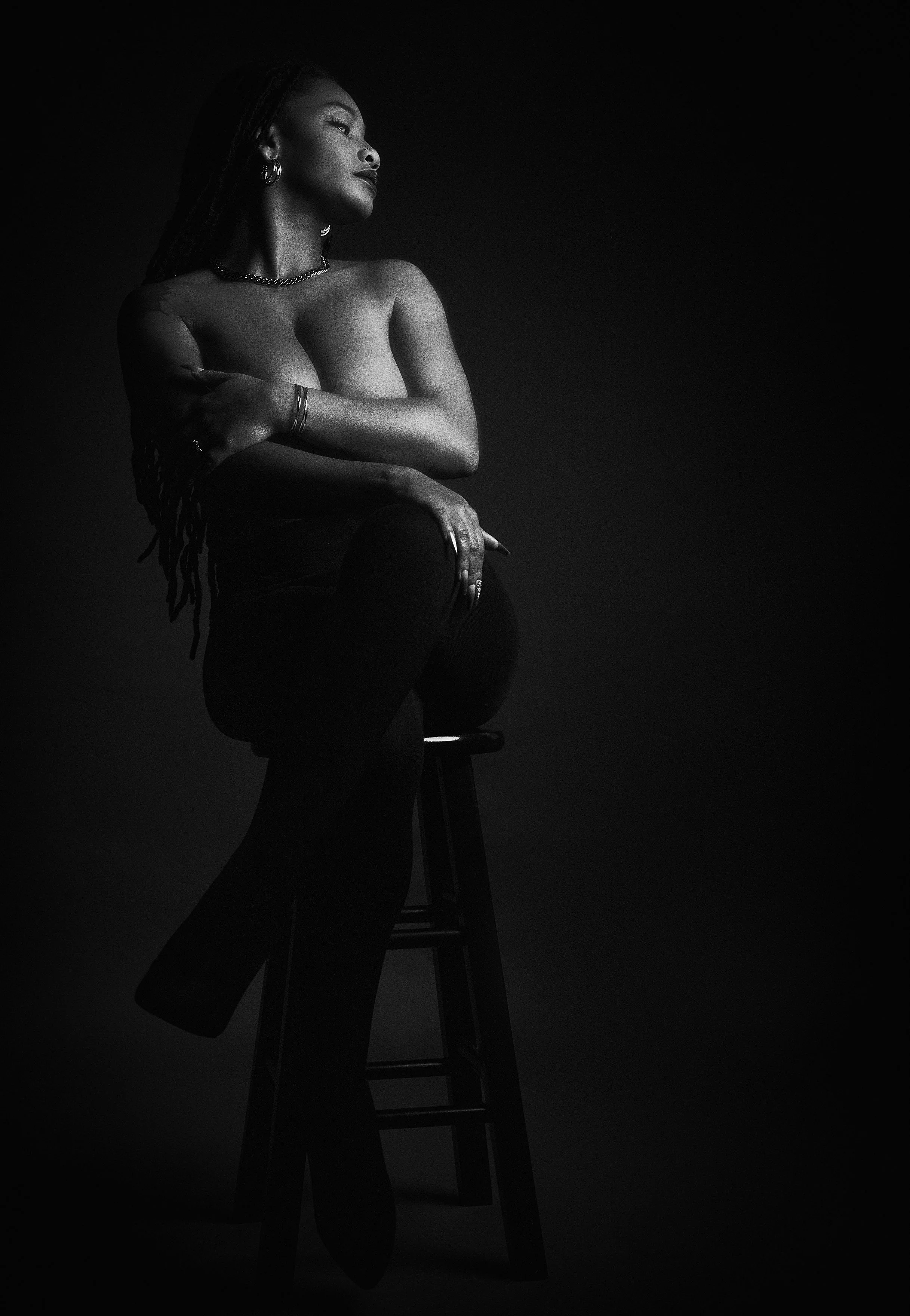 Black and white photo of a woman sitting on a stool, topless, with her arms crossed over her chest, looking to her left against a dark background.