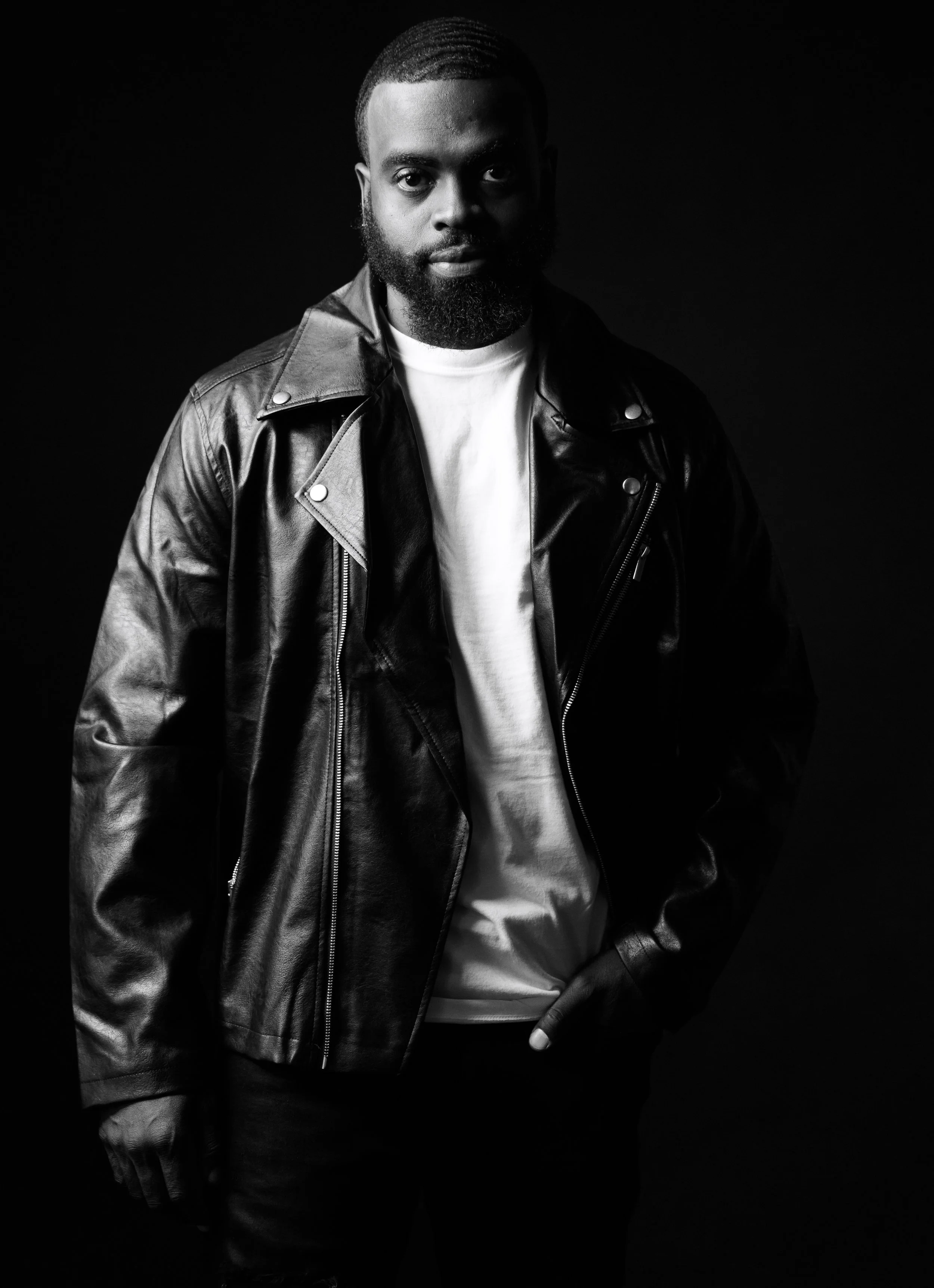 Black and white photo of a man with a beard wearing a leather jacket and white T-shirt against a dark background.