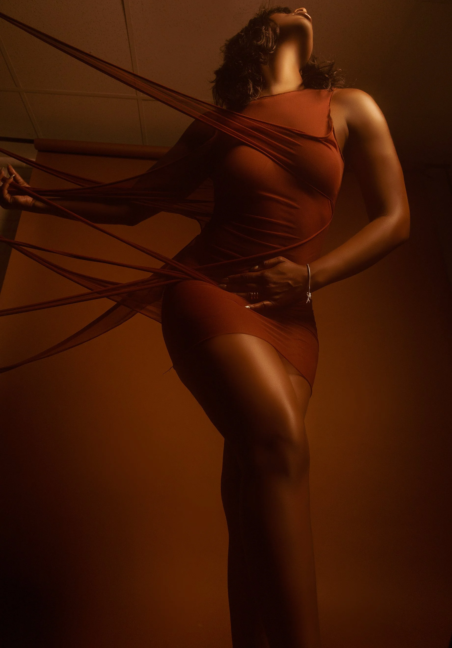 A woman in a sheer, tight red dress posing with her face tilted upward and her hand on her thigh, with fabric streams extending from her arm in a dramatic, artistic fashion.