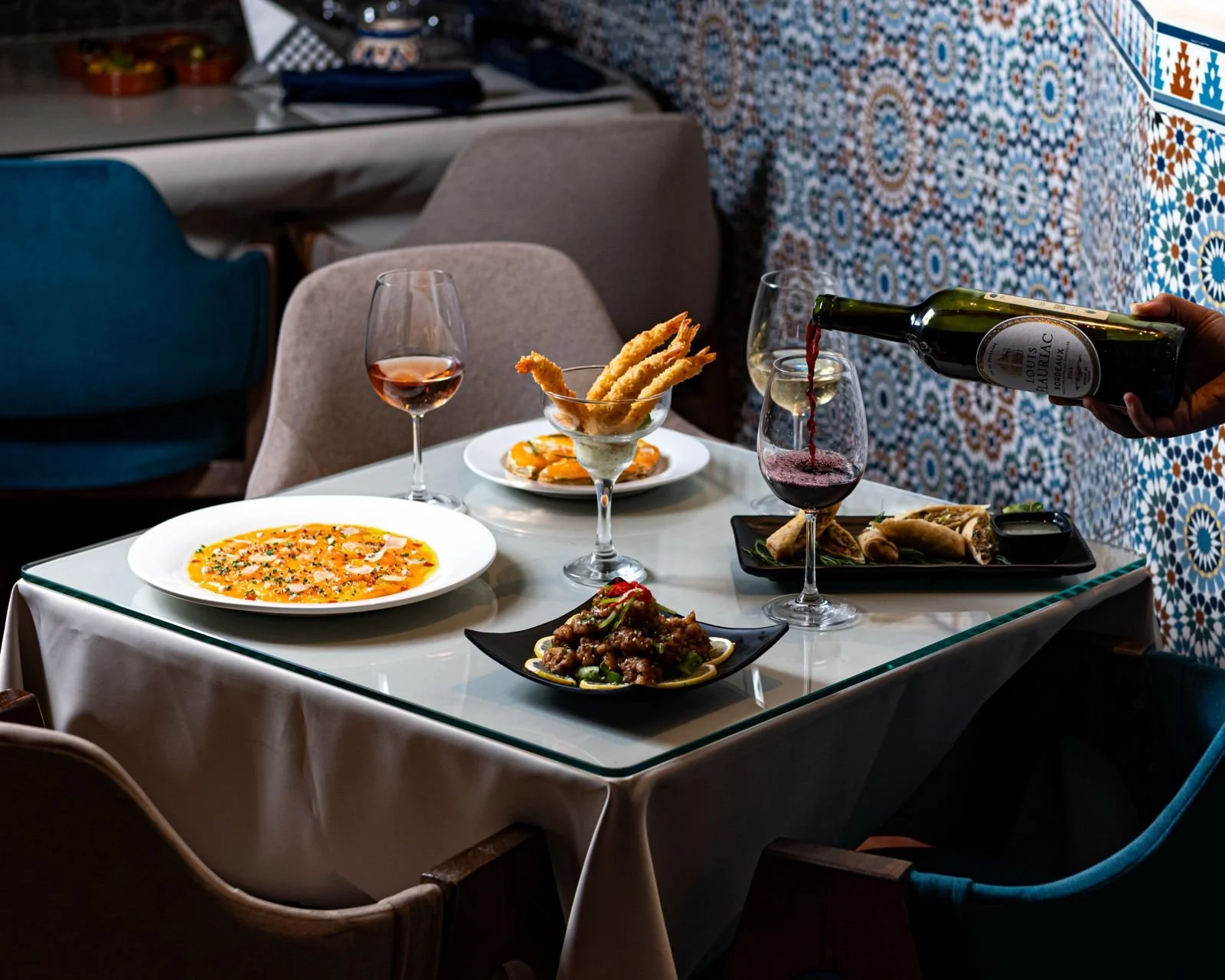 Une table de restaurant avec divers plats et deux verres de vin, un spectateur verse du vin rouge dans un verre.