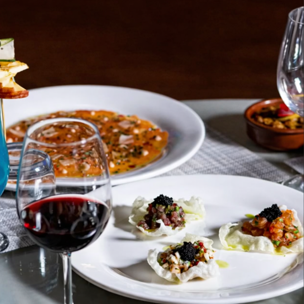 Assiette avec trois petites portions de tartare de poisson garnies de caviar noir, verre de vin rouge et plat en arrière-plan avec une tartine à l'airrouge