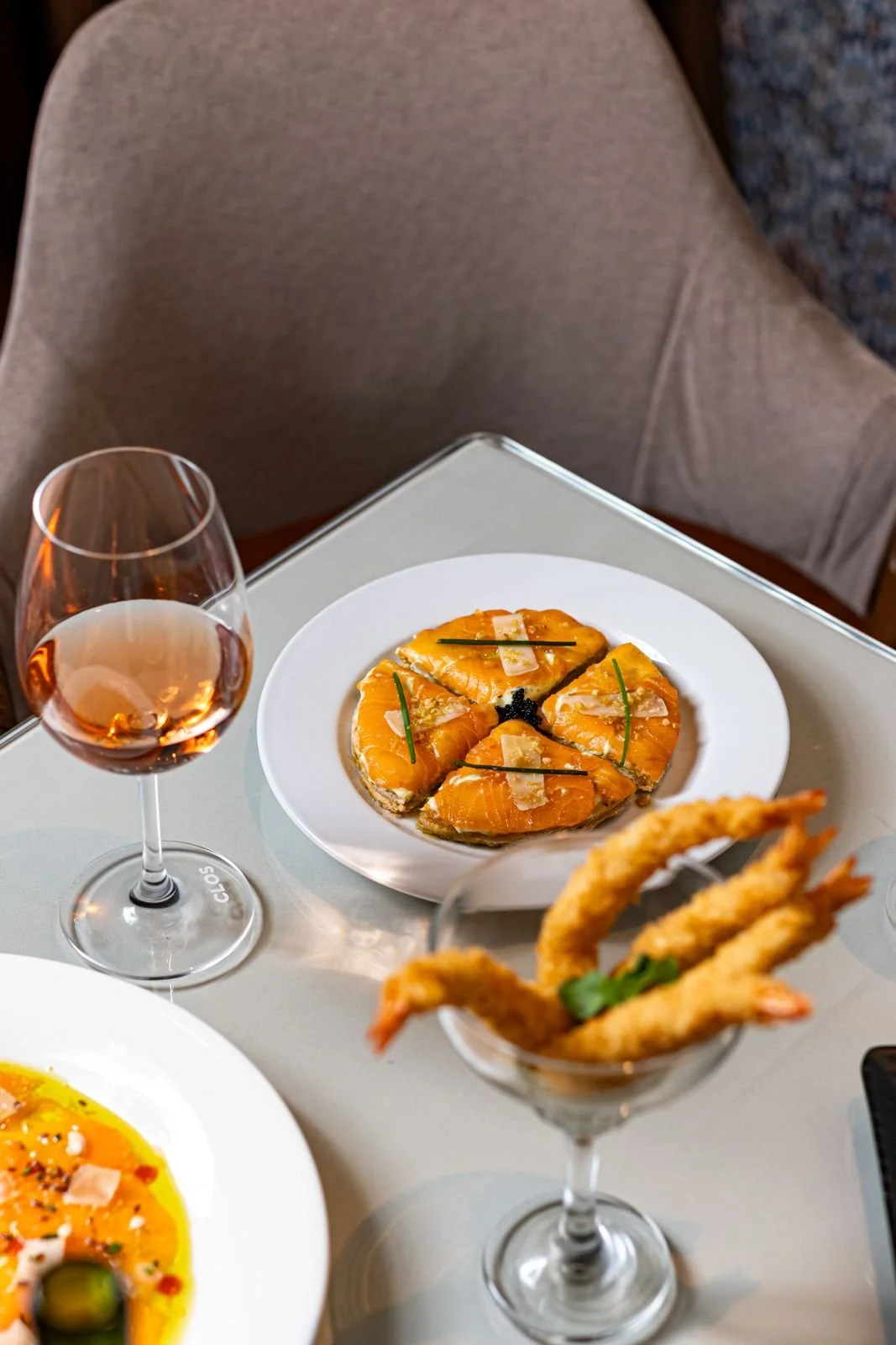 Assiette de tapas avec des tranches de saumon garnies de truffes et d'herbes, verre de vin rosé, et une coupe contenant des crevettes frites, sur une table à manger.
