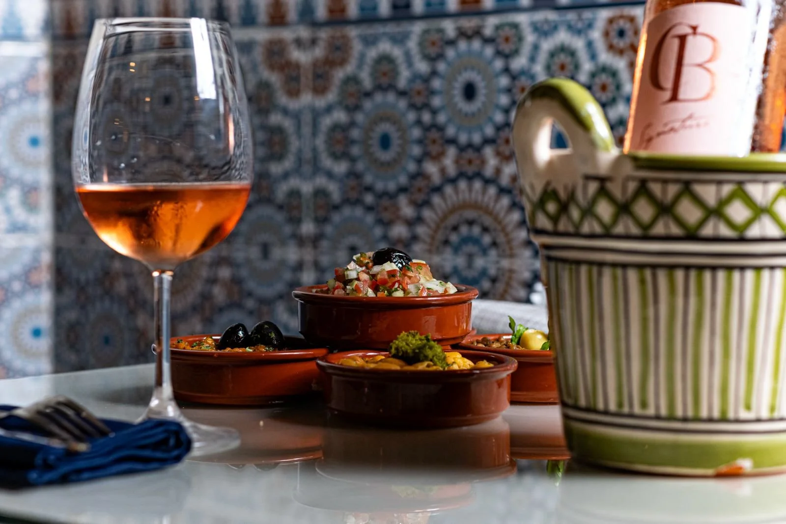 Verre de rosé, plats de nourriture méditerranéenne dans des plats en terre cuite, bol de salade de légumes, assiette de légumes et de fromage, panier à pain avec une bouteille de rosé, fond coloré avec motif géométrique.