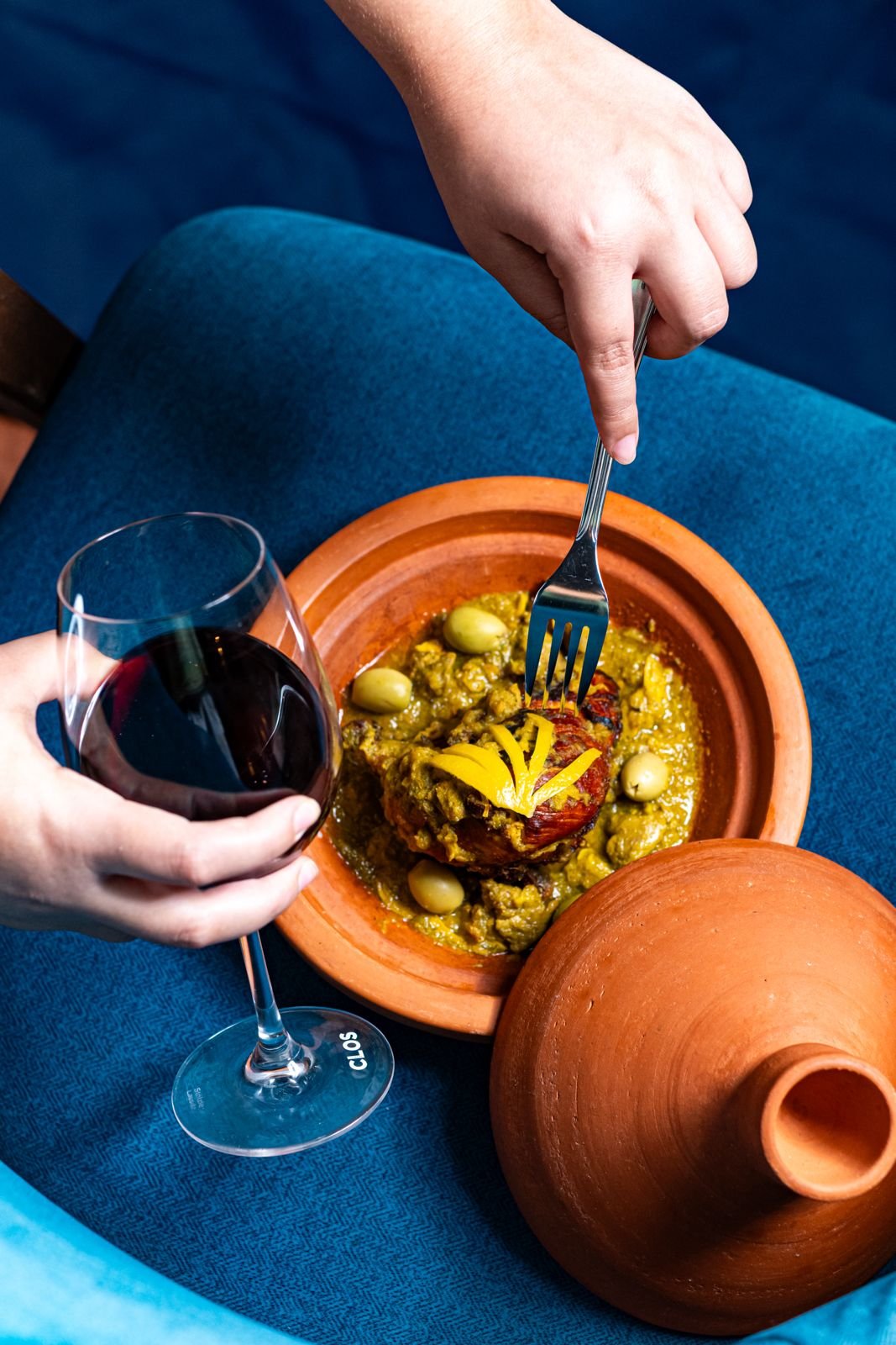 Une personne tient un verre de vin rouge dans une main, tandis qu'une autre personne utilise une fourchette pour couper un plat traditionnel dans un bol en terre cuite, avec un jarre en terre cuite à côté.