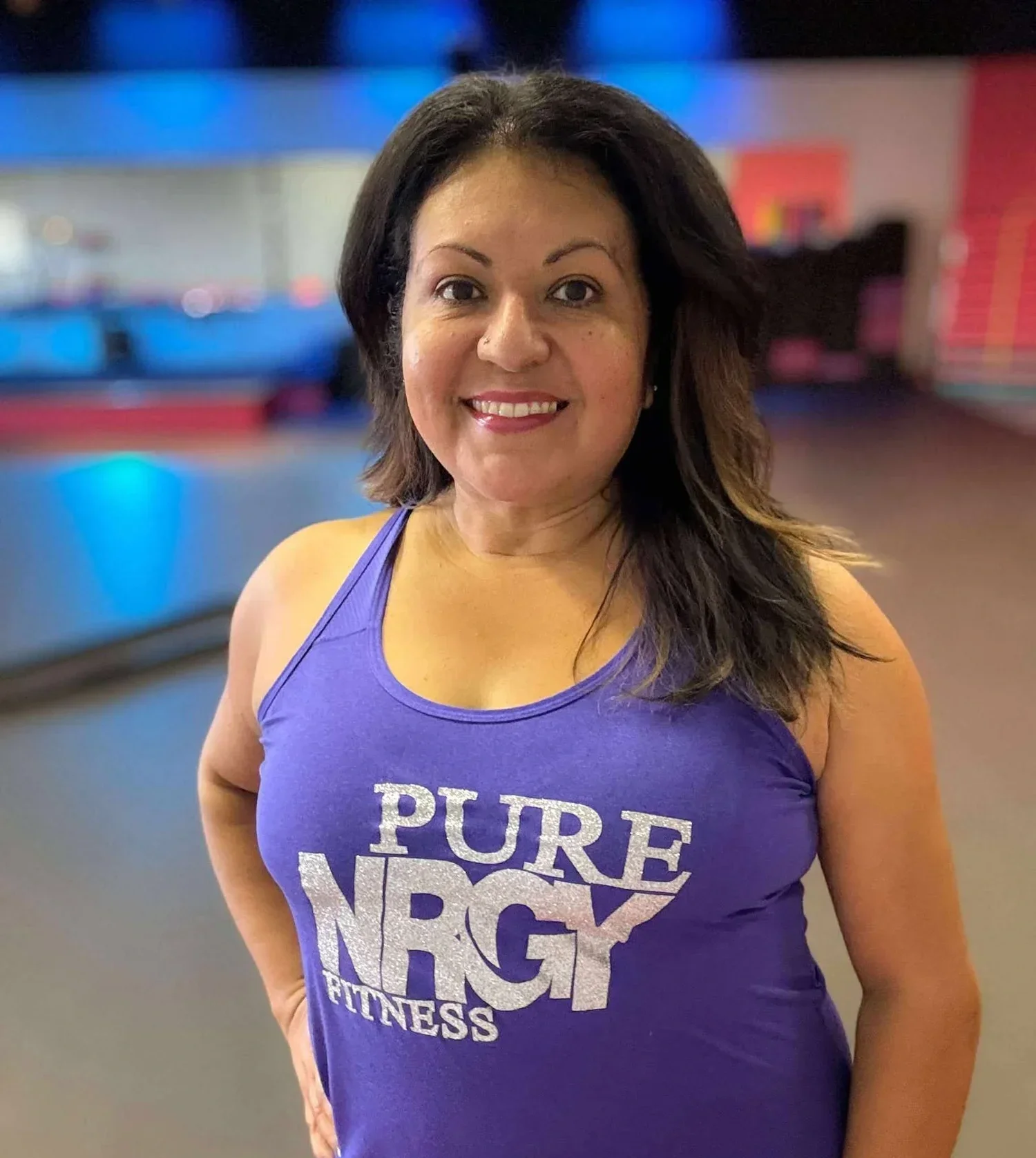 Woman smiling wearing a purple tank top with the text 'Pure NRG Fitness' on it, in a gym setting with blurred background.
