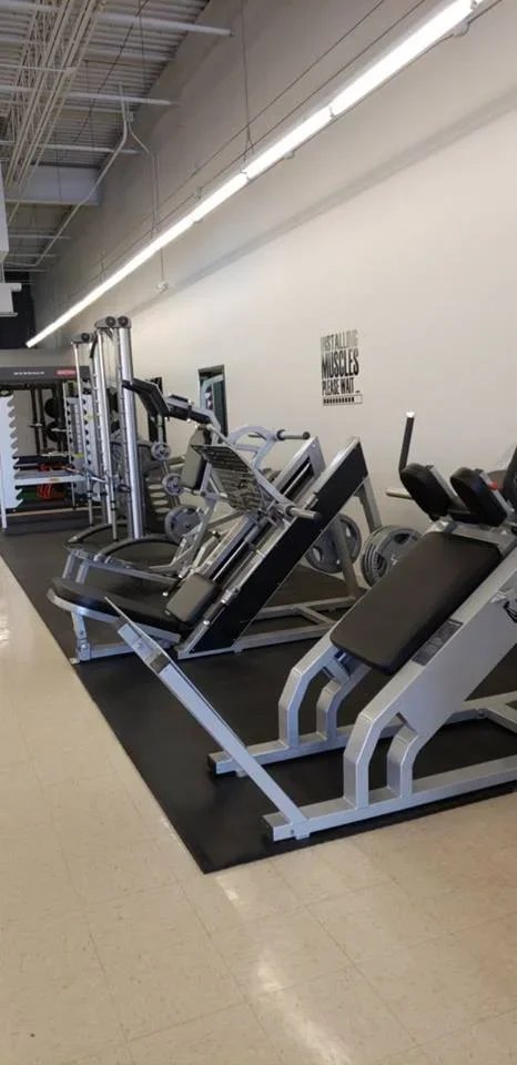 Gym equipment including leg press and exercise machines along a white wall with a motivational quote, in a well-lit workout space.