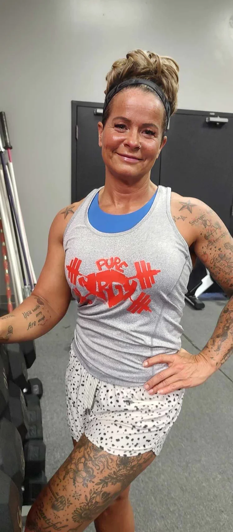A woman with tattoos, wearing a tank top with the words 'PURE HARD' and a barbell design, in a gym. She has a headband, short shorts, and is smiling at the camera.