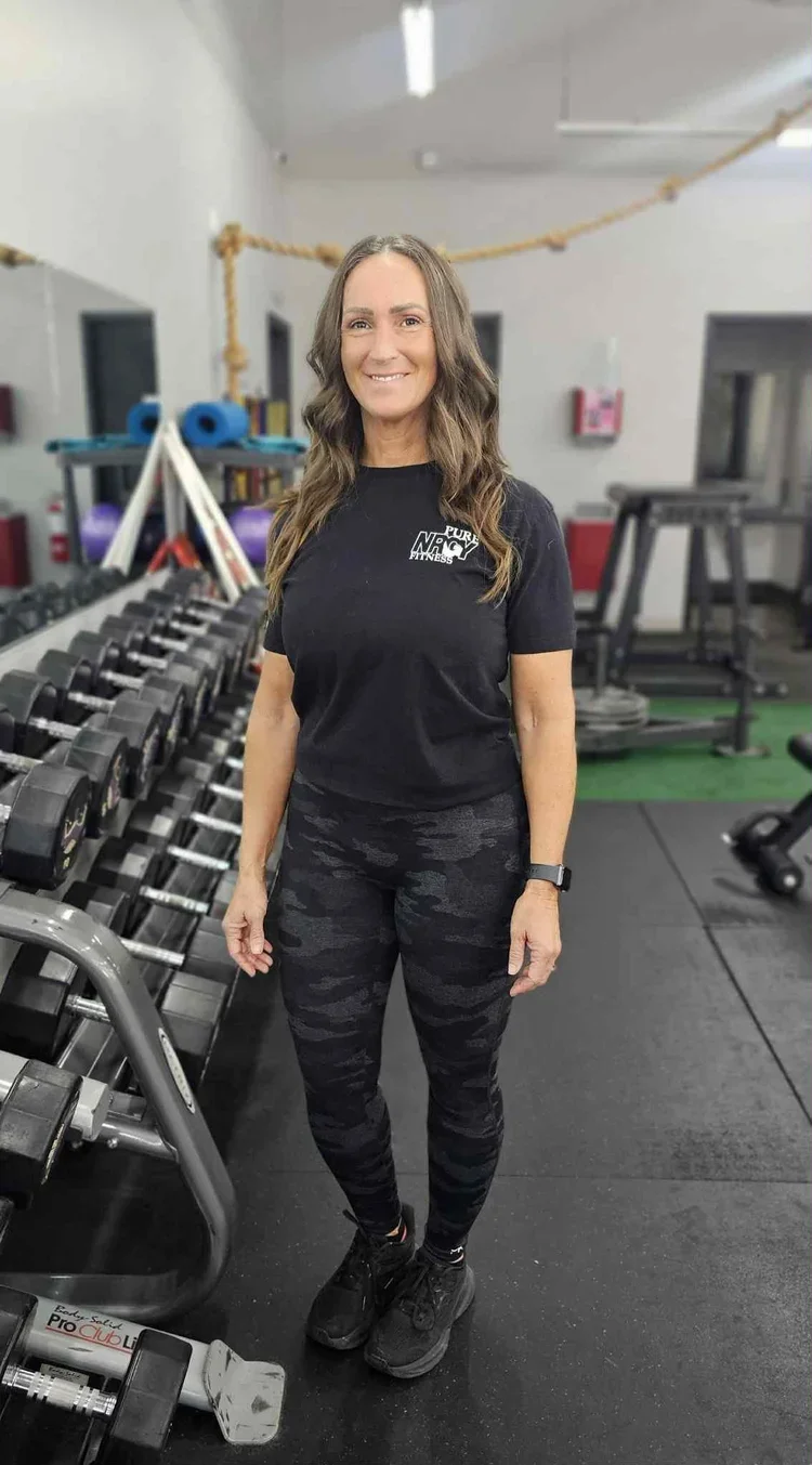 A woman with long brown hair smiling in a gym, wearing a black t-shirt, black camouflage leggings, and black athletic shoes, standing next to dumbbells and workout equipment.