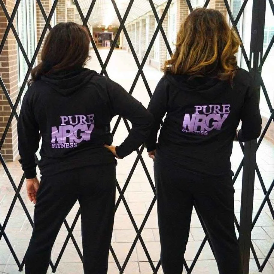 Two women wearing black hoodies with purple and white 'Pure NRG Fitness' logos on the back, standing side by side behind a black gate, outside a building with brick walls and large windows.