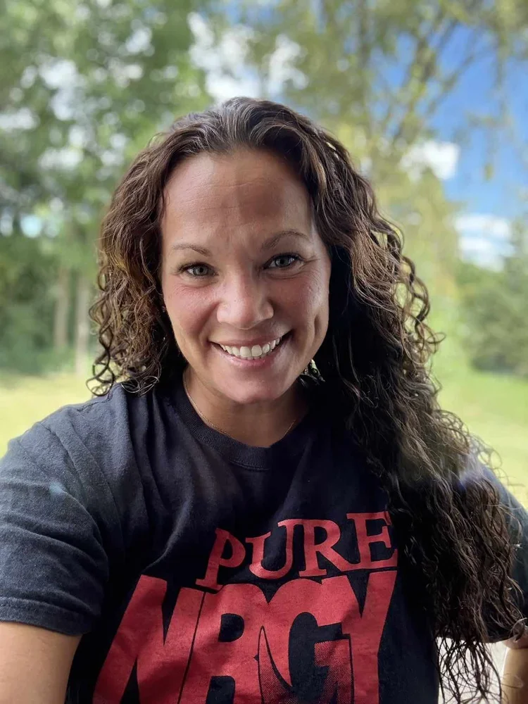 A woman with long curly brown hair and a tan complexion smiling outdoors with a blurry background of green trees and blue sky. She is wearing a black T-shirt with red text that reads "PURE BRAV".